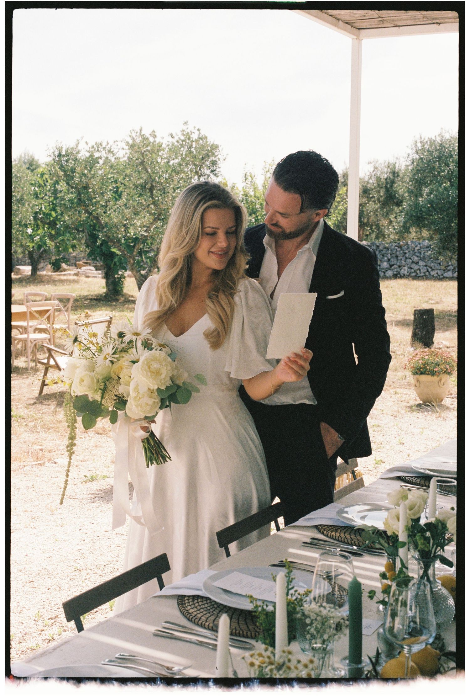 Film photo of a bride and groom reviewing wedding vows at an outdoor table in an olive grove.
