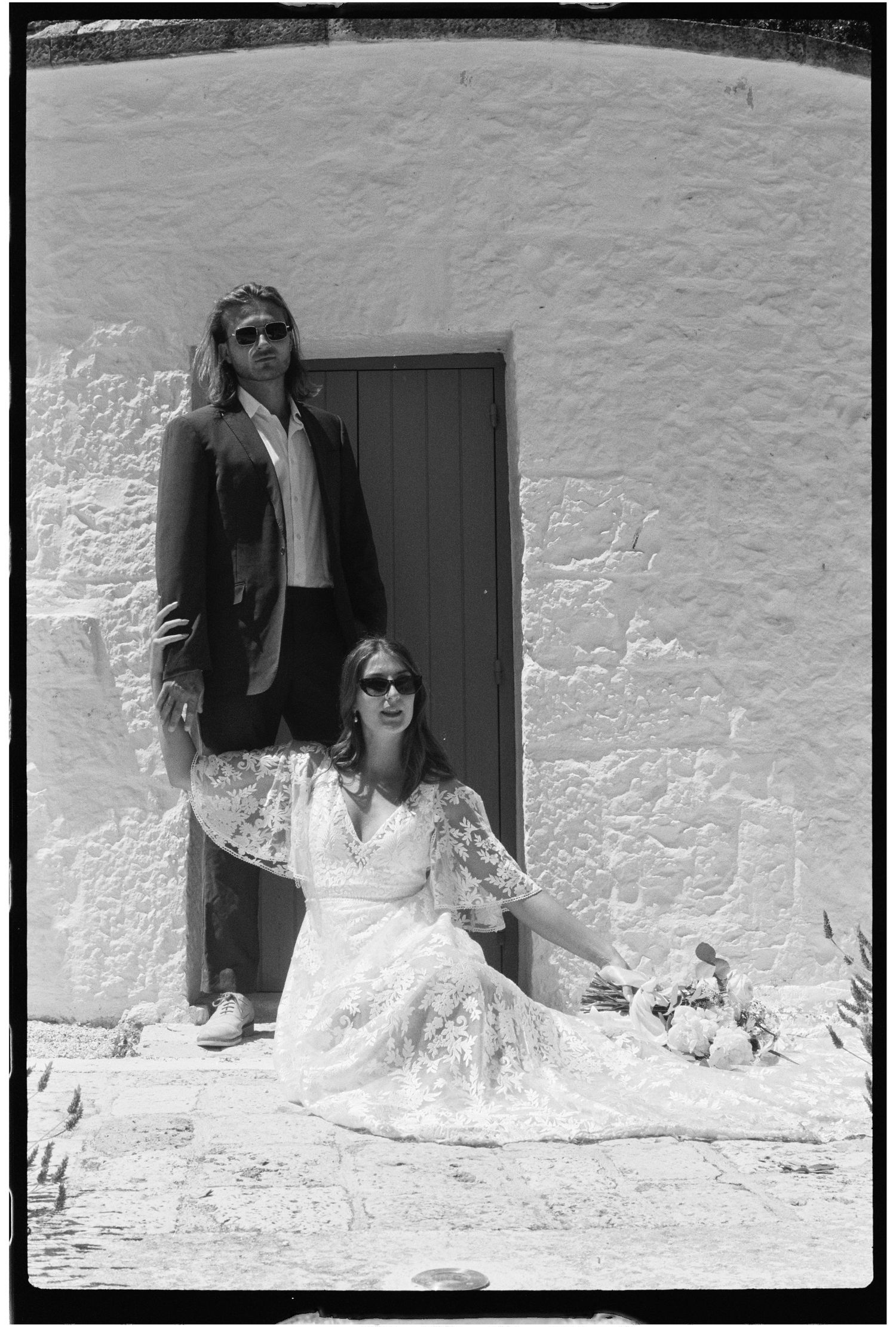 Black and white photo of a couple posing by a white wall, woman in lace dress seated, man standing in dark jacket.