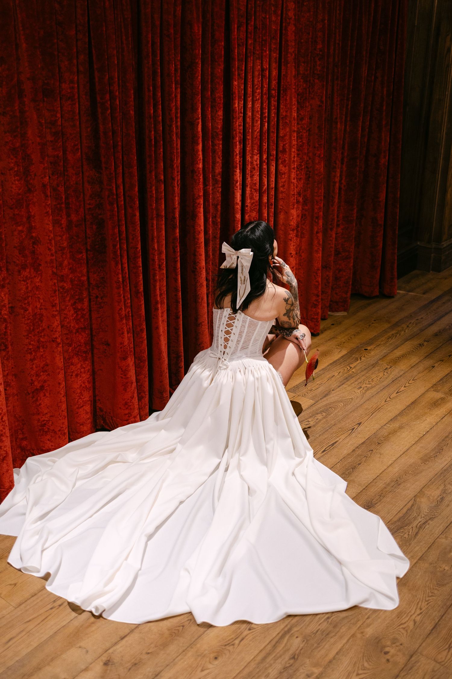 Bride in white satin ballgown with corset back and bow, seated on wooden floor before red velvet curtains.
