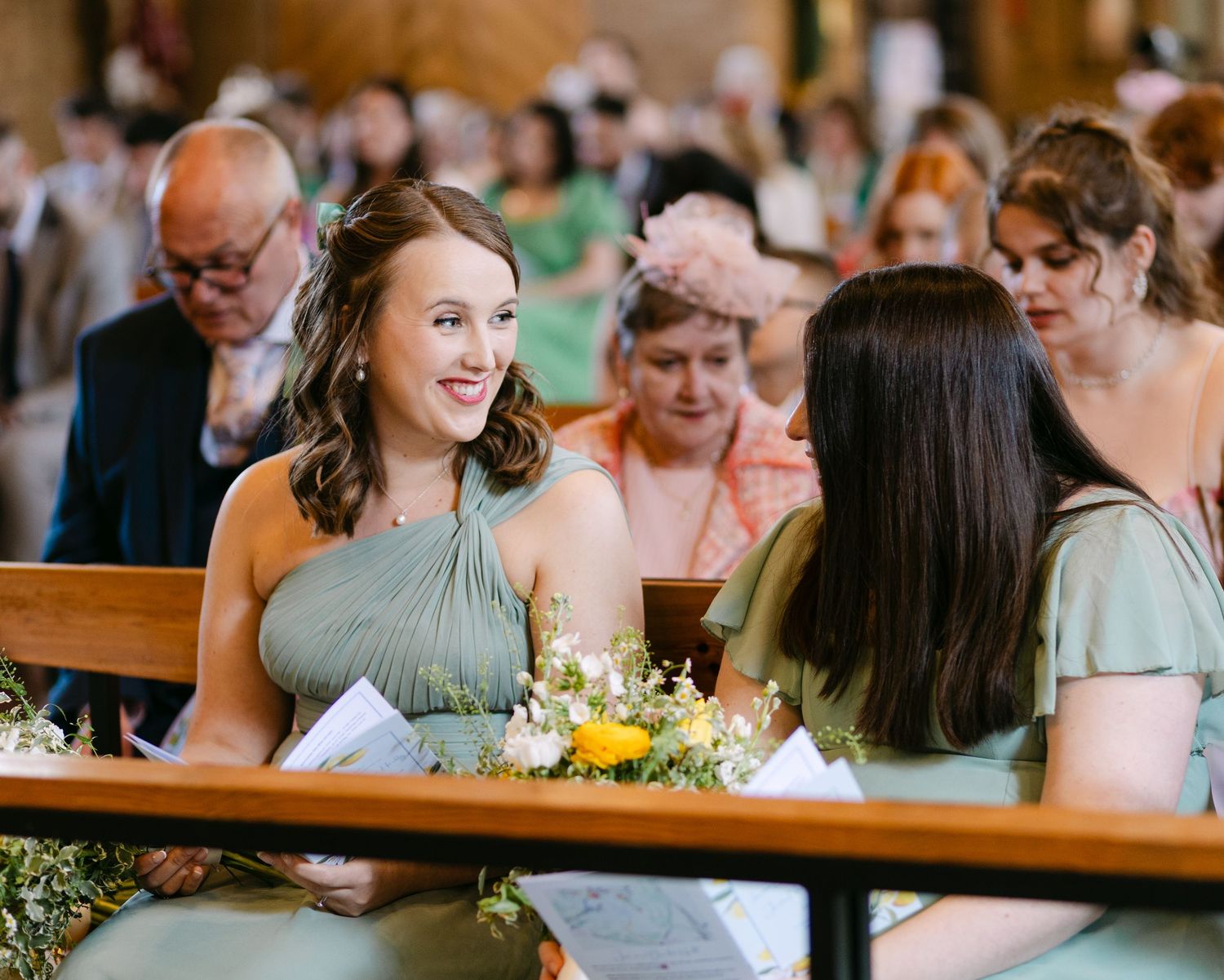 Smiling bridesmaids in sage green dresses chatting inside a church before the wedding ceremony begins.