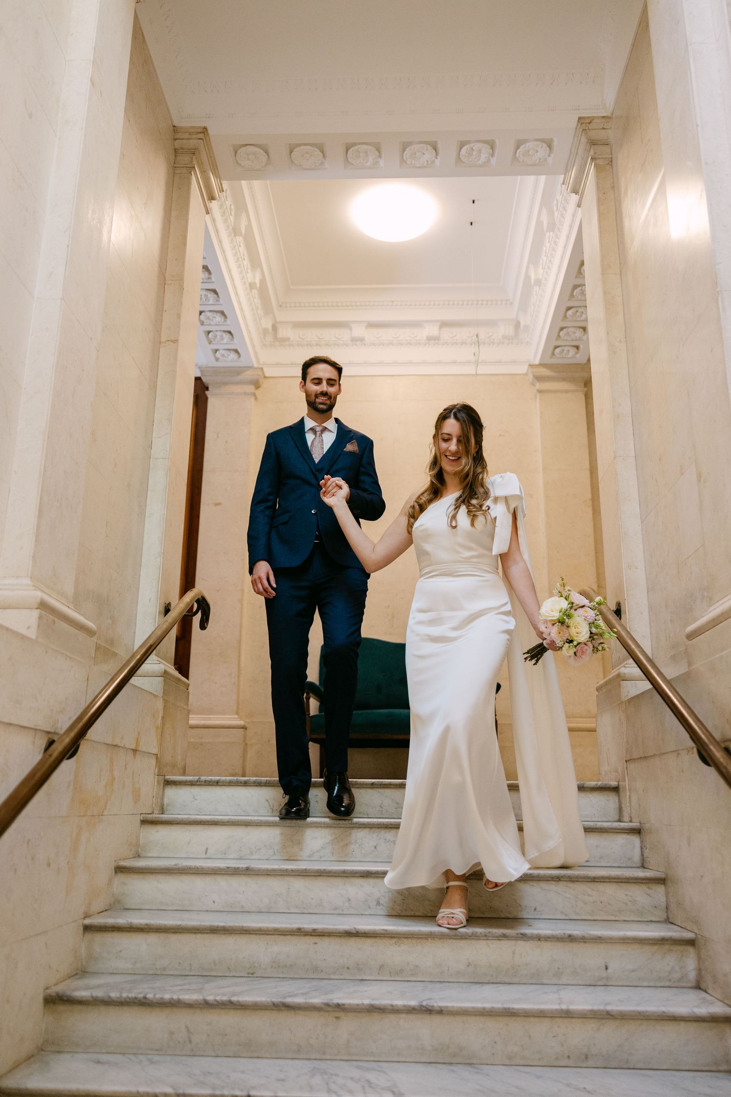 A couple descends elegant white marble stairs in formal wedding attire.