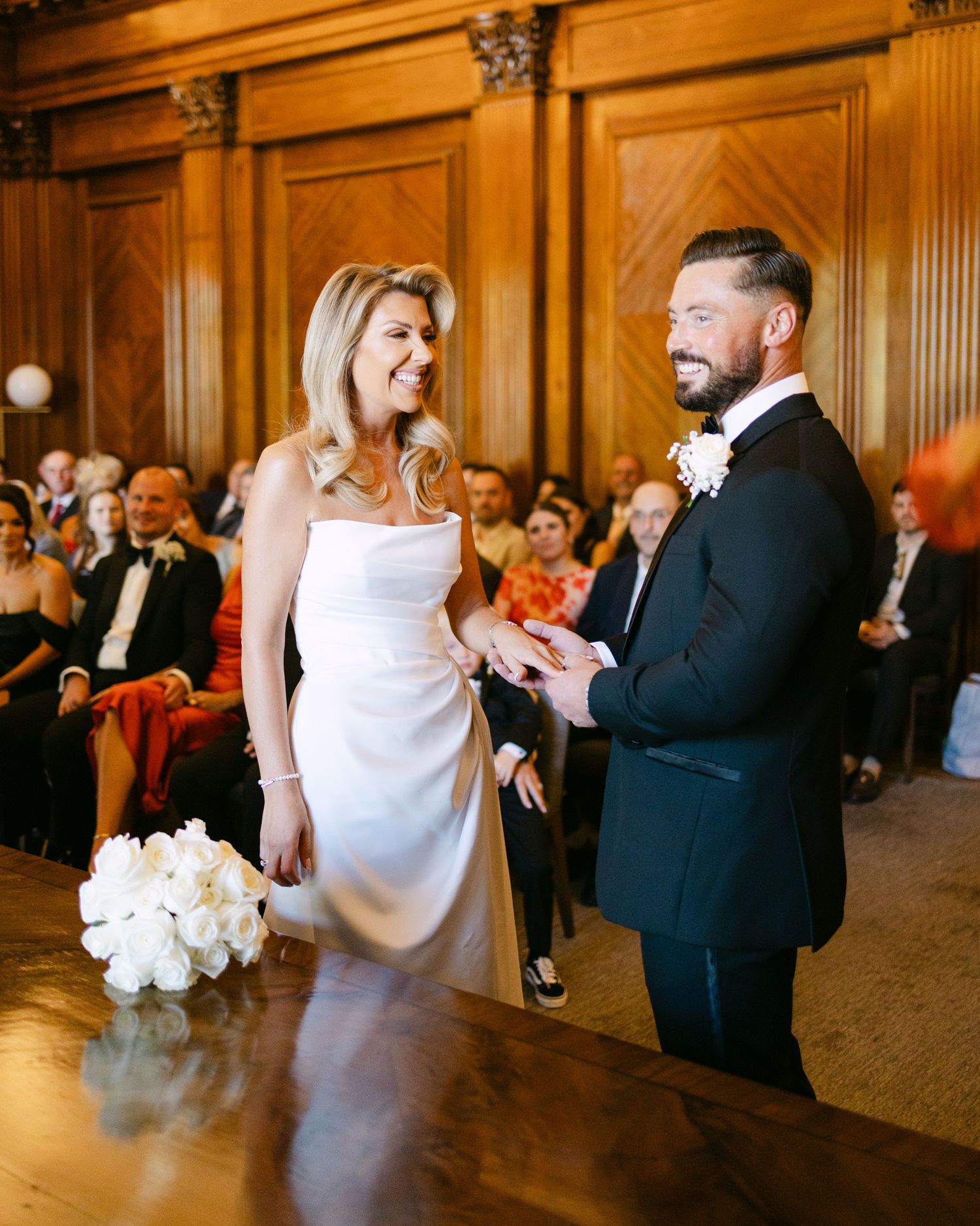 A wedding ceremony taking place in an elegant wood-paneled room with guests seated in red chairs.
