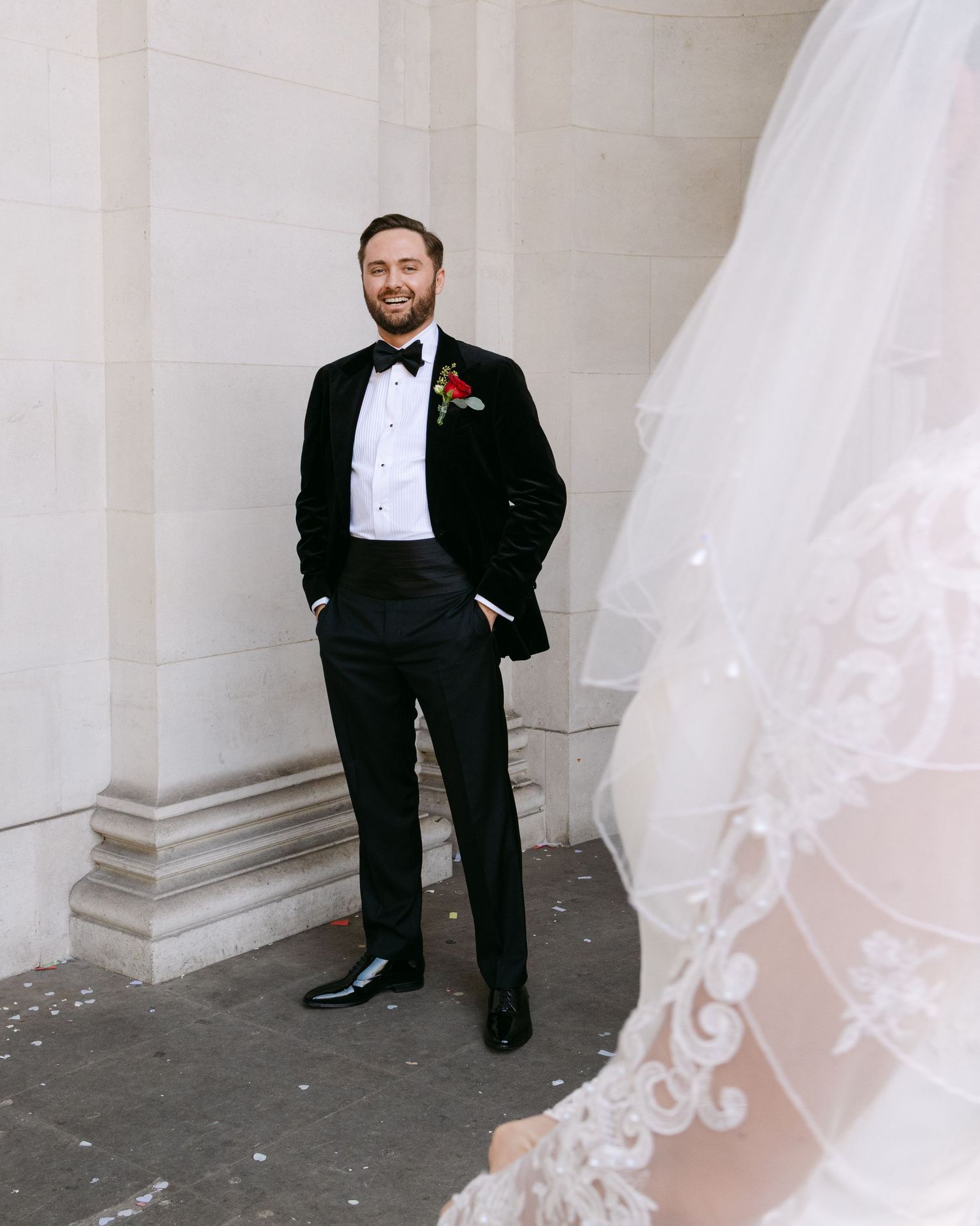 Elegant groom in black tuxedo poses against white pillared wall with bride's veil visible.