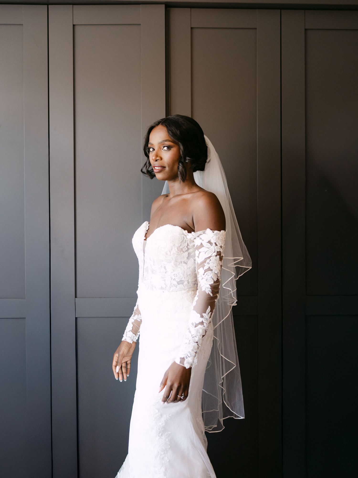 Elegant bridal portrait showing off detailed lace sleeve wedding dress against dark grey wall.