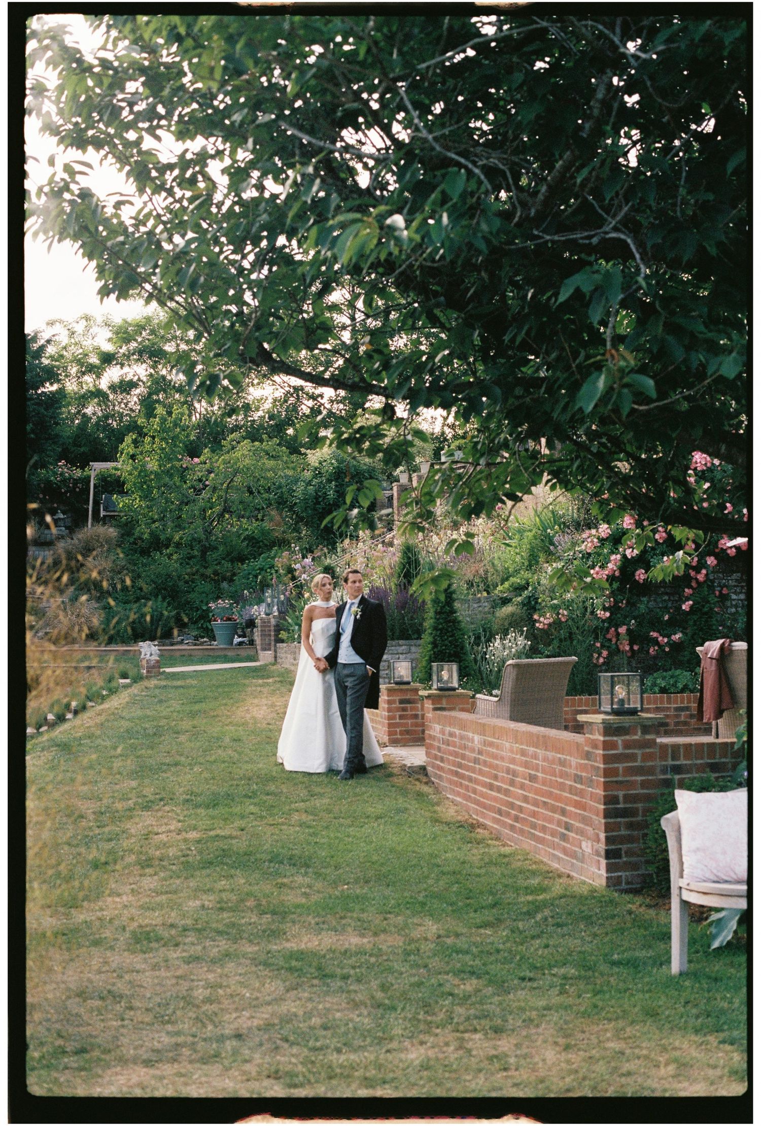 Bride and groom standing together in a lush garden beside a brick wall at dusk.