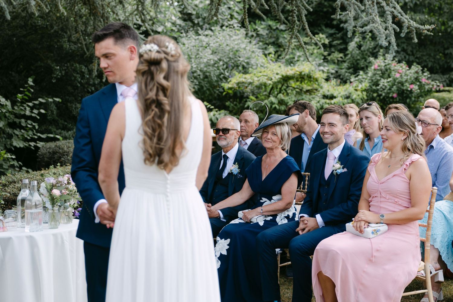 Outdoor wedding ceremony with bride and groom facing smiling guests seated in garden chairs amid lush greenery.