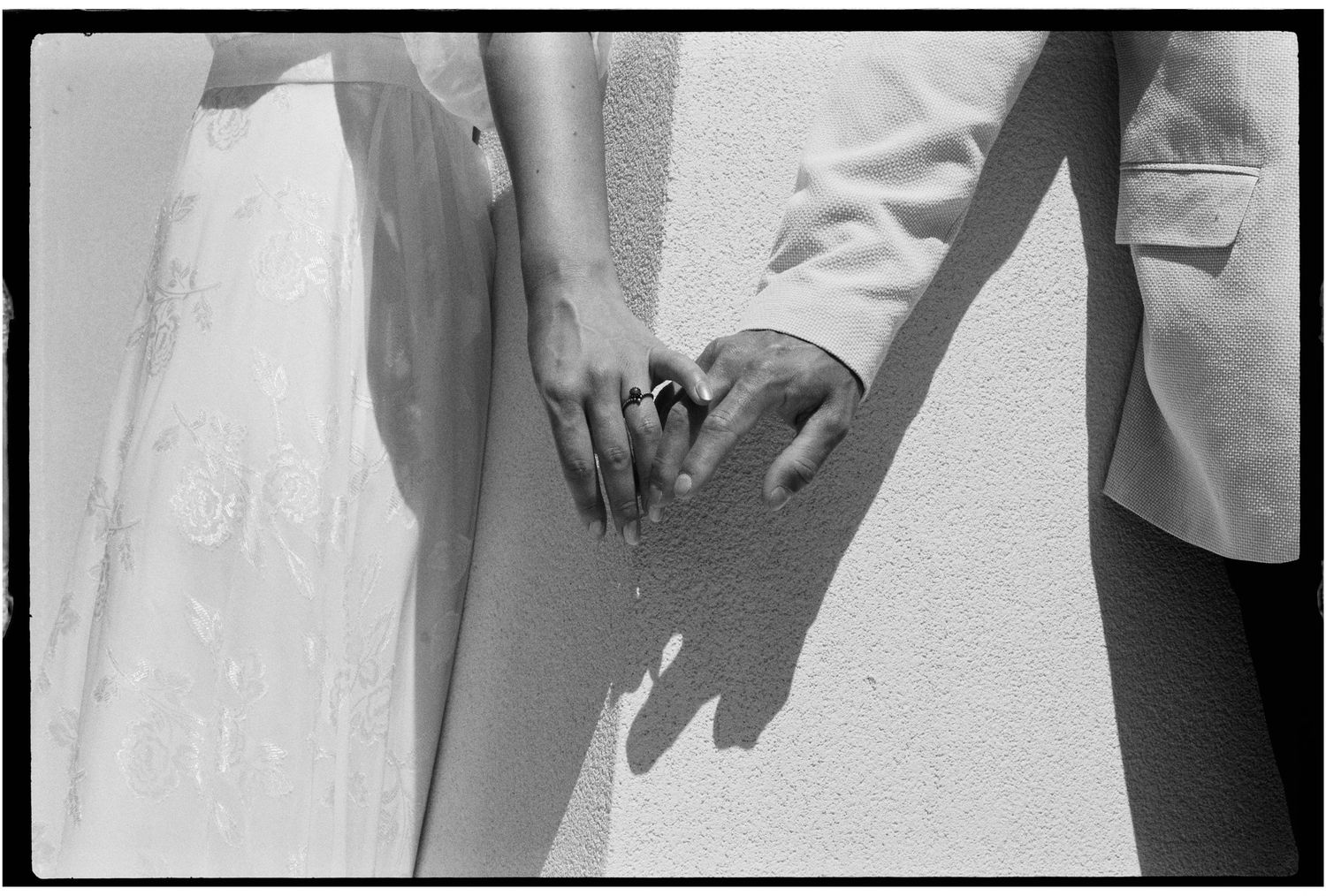 Black and white photo of a bride and groom holding hands, showing wedding rings and formal attire.
