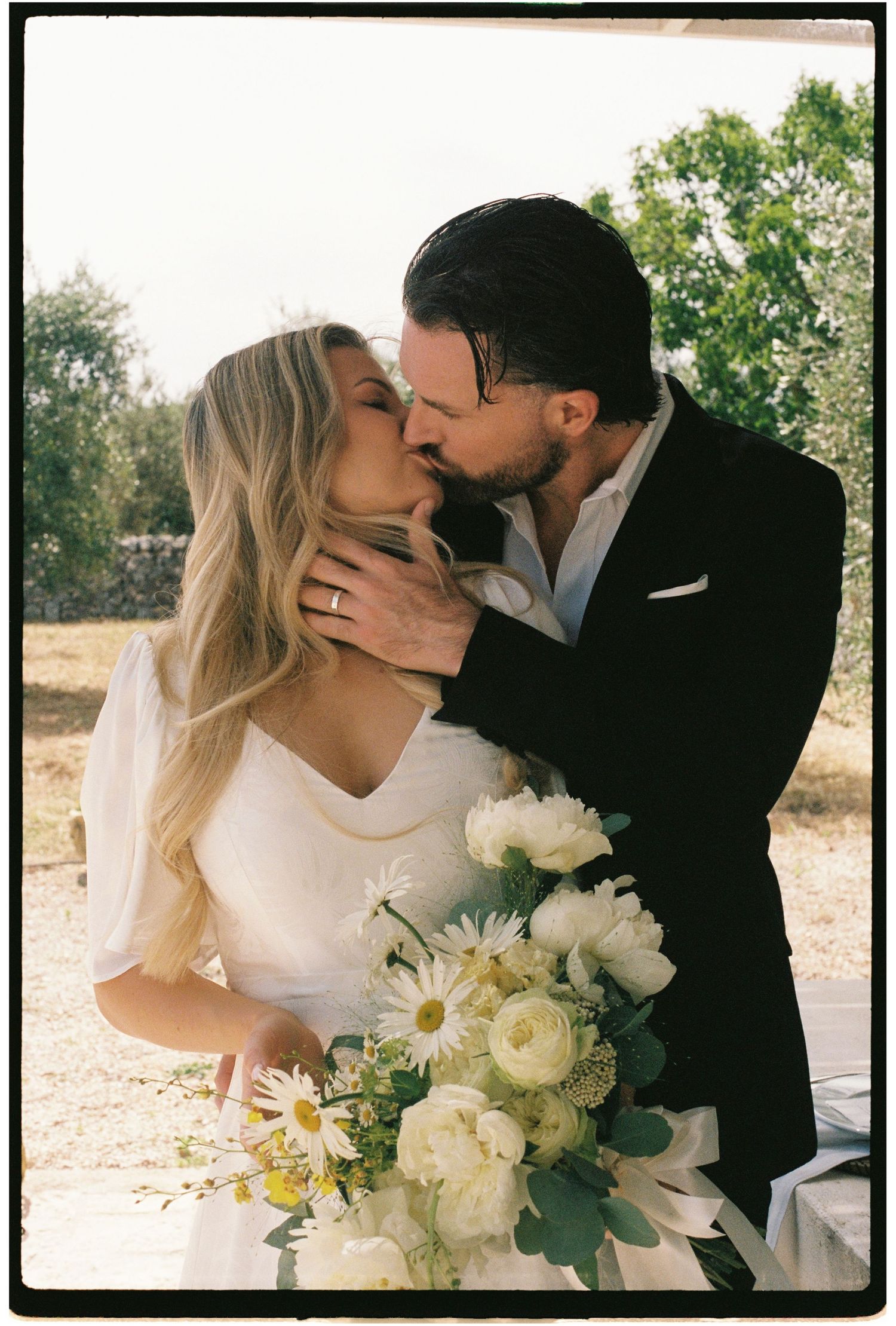 Couple sharing a kiss at their outdoor wedding, bride holding white floral bouquet.