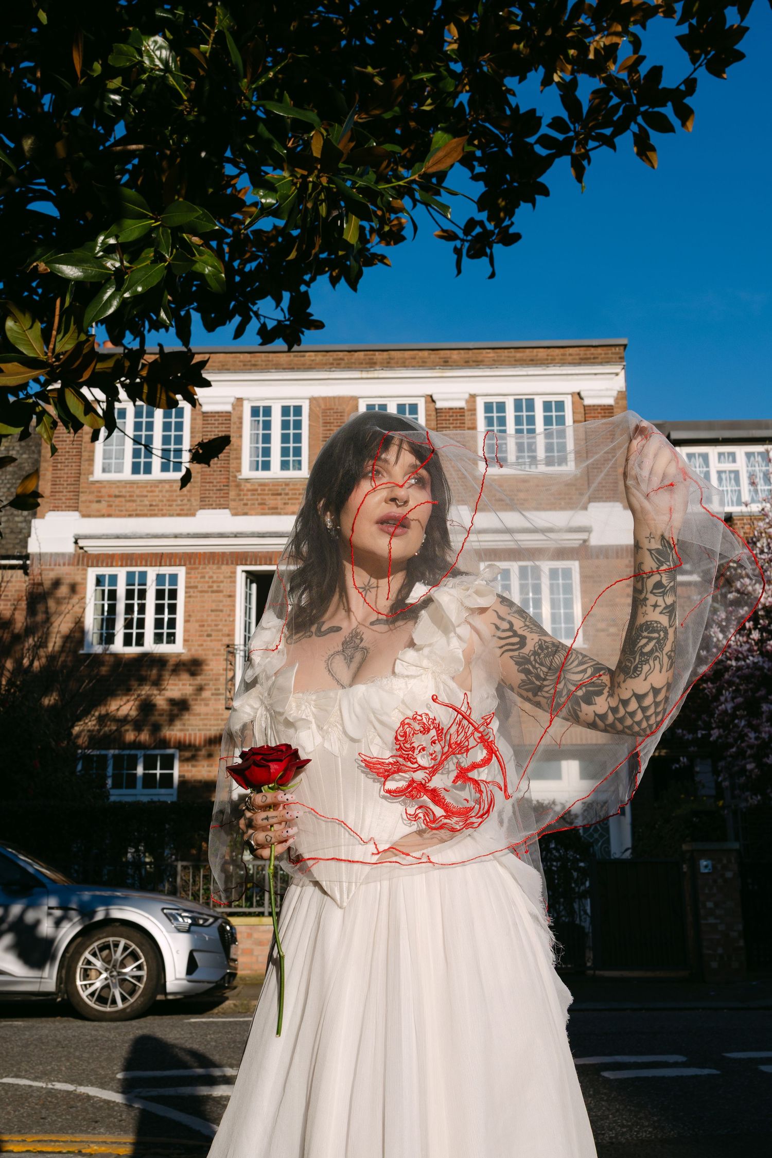 Tattooed bride in white gown with red cherub veil holds a rose outdoors near a brick building on a sunny day.