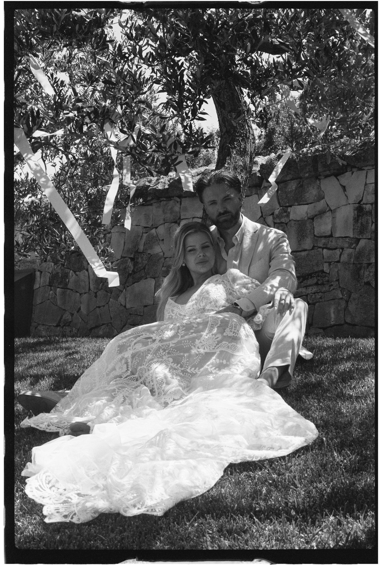 Bride and groom sit together on grass beneath a tree with paper streamers floating in the air.