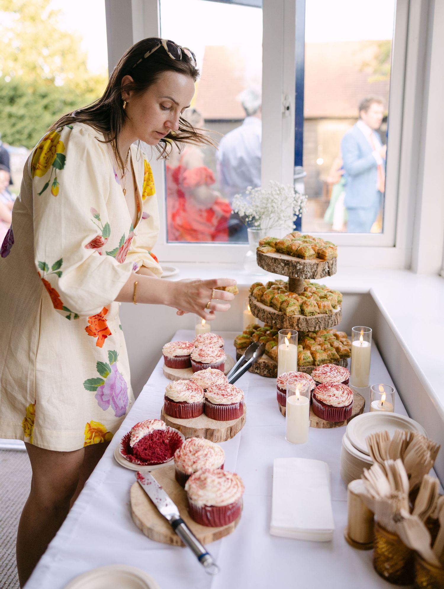 A floral-dressed figure arranges pink frosted cupcakes and desserts on a white tablecloth at an event.