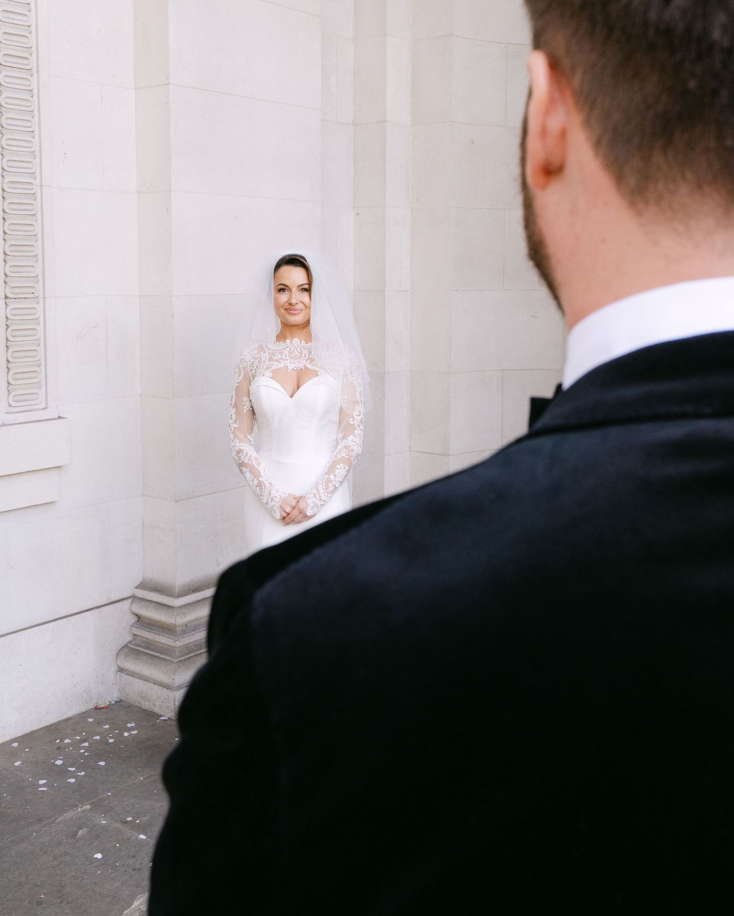 Bride in a white lace wedding dress poses against a light stone wall for artistic wedding photos.