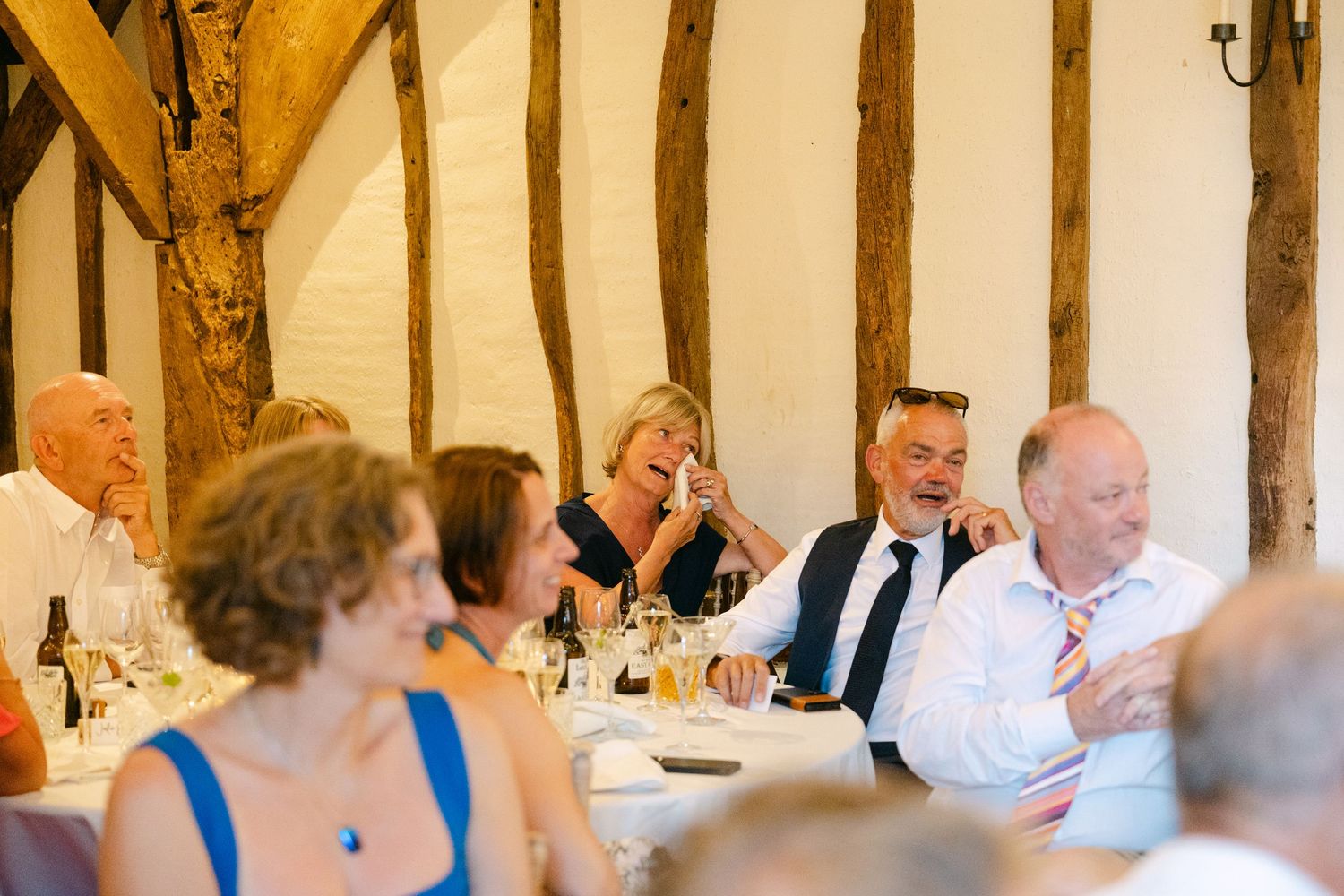 Wedding guests laughing and enjoying speeches at a rustic barn reception venue with timber beams.