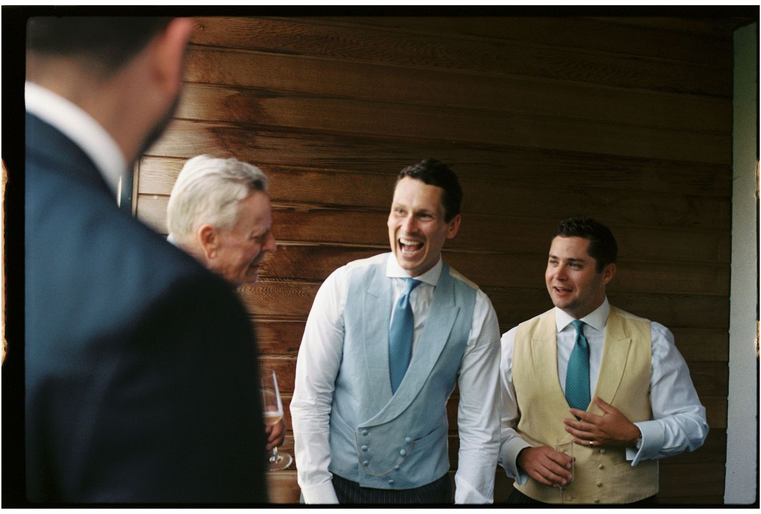 Three men in formal attire laughing together at a wedding celebration near a rustic wooden wall.