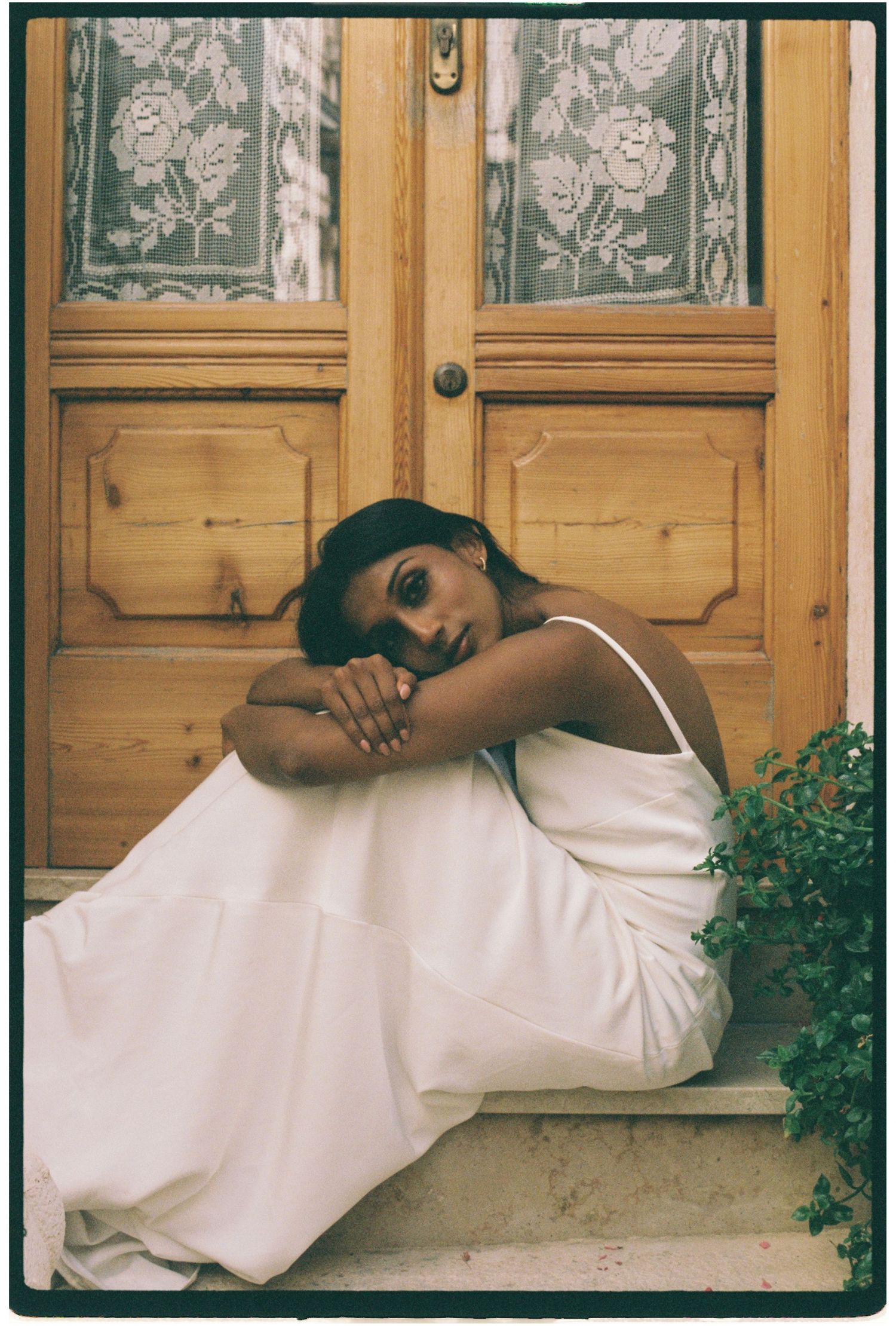 Woman in white slip dress sitting on steps in front of a wooden door with lace curtains.