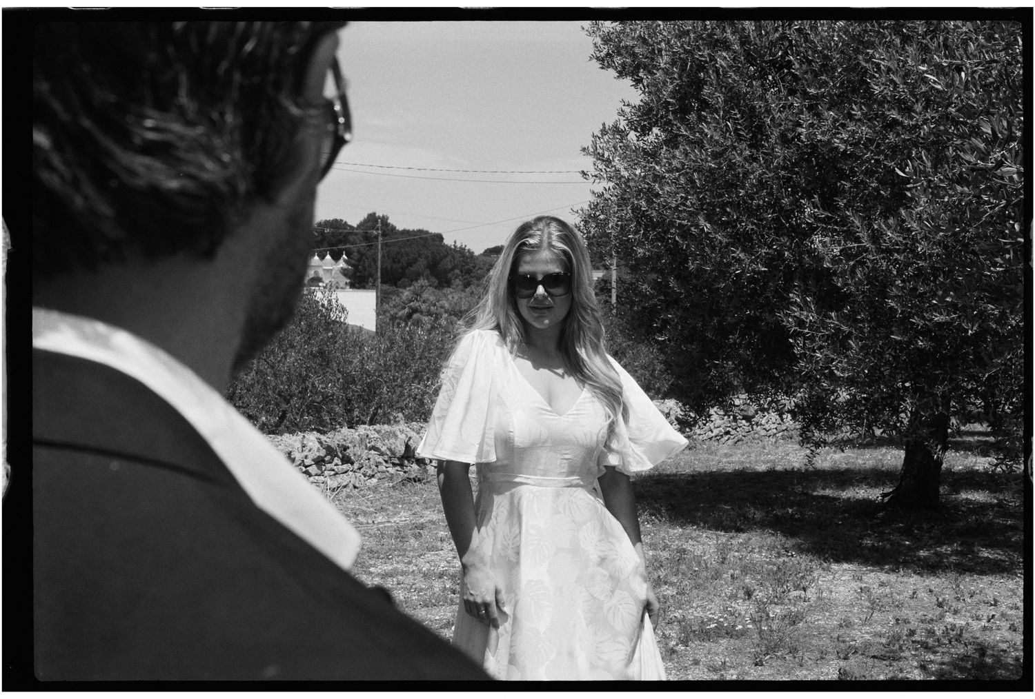 Black and white photo of a bride in white dress smiling outdoors near olive trees at a wedding.