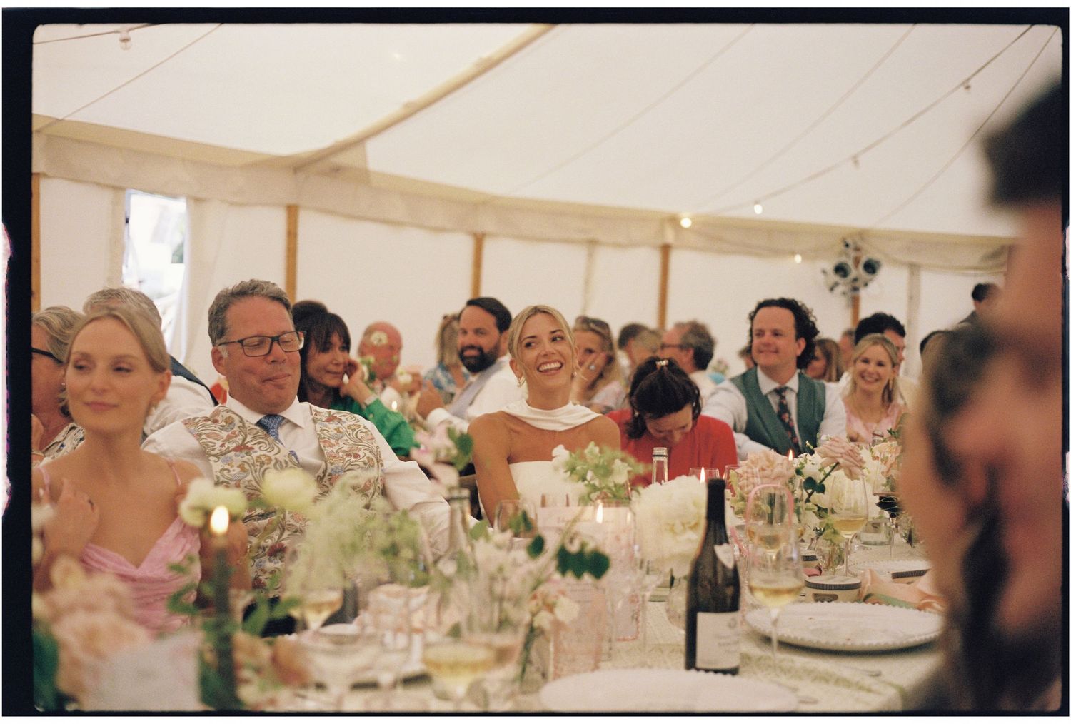 Film photo of a smiling bride seated at a long decorated table during a wedding reception in a marquee.