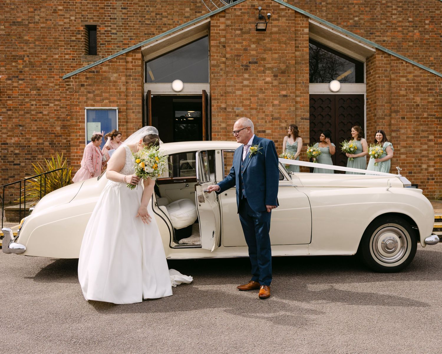 Bride arriving at church in a classic white Rolls-Royce, assisted by a man in a blue suit, with bridesmaids in background.