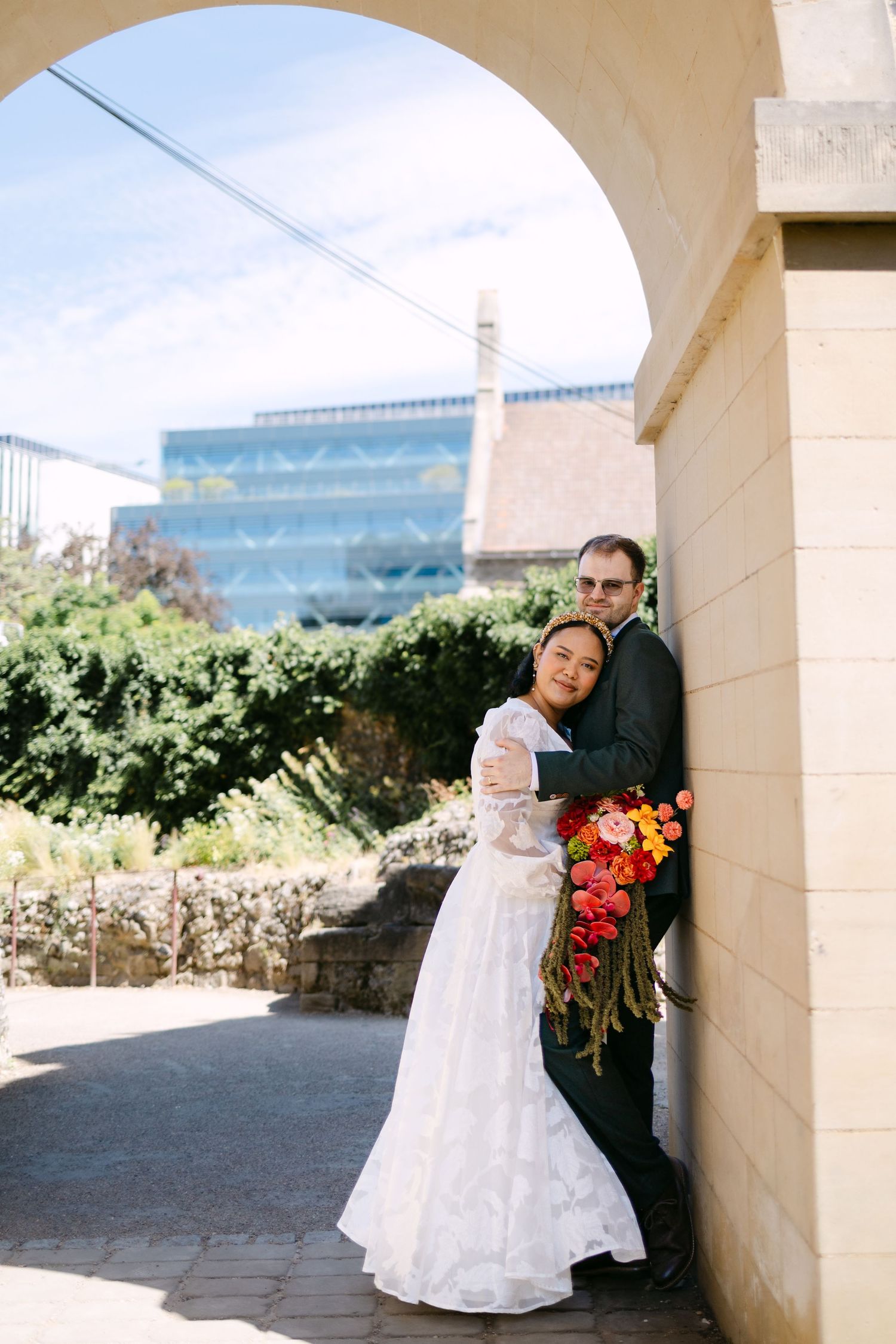 Bride and groom embracing under stone arch on wedding day, bride holding red bouquet, outdoor venue with blue building.