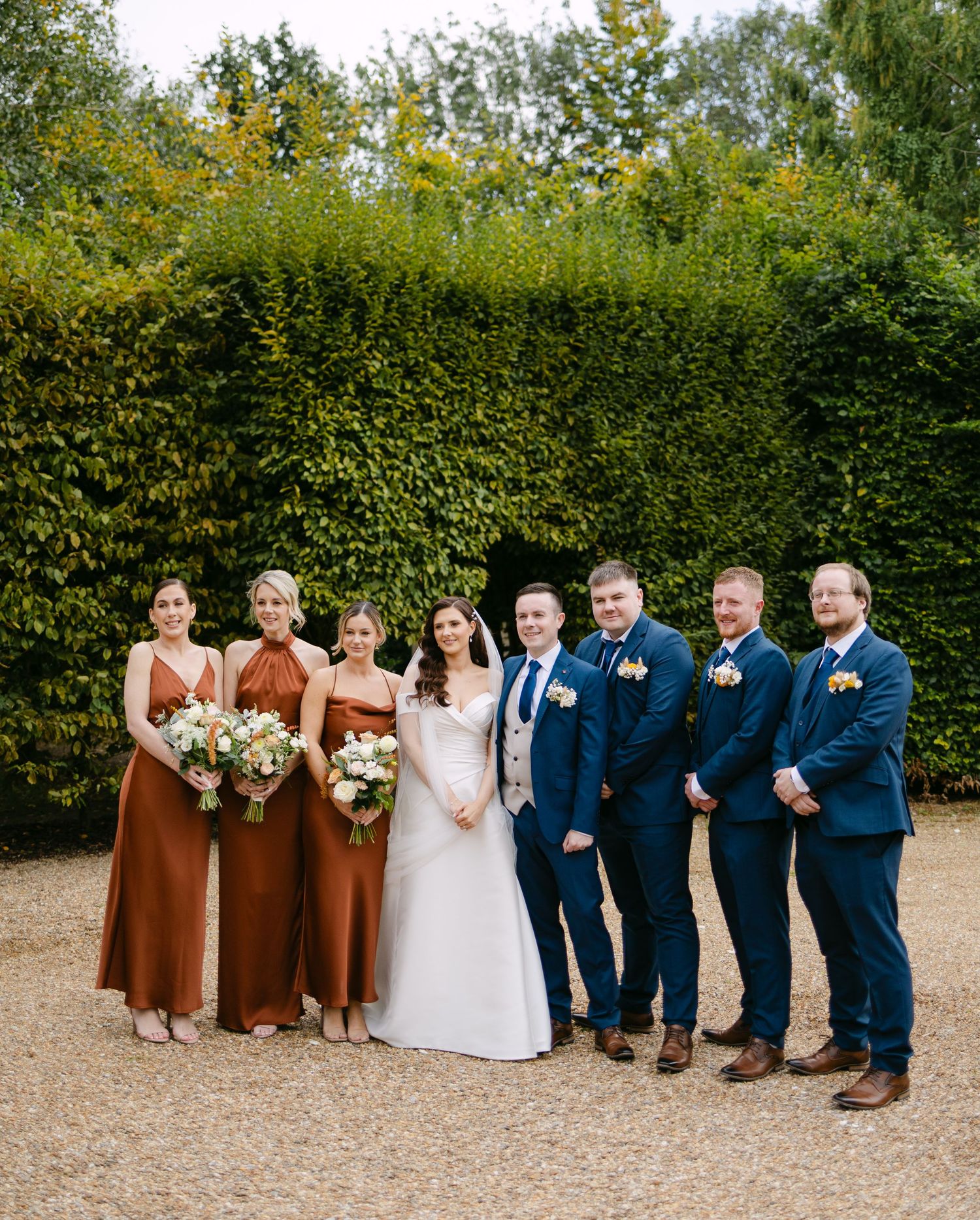 Wedding party in navy suits and rust-colored dresses poses for formal photos.