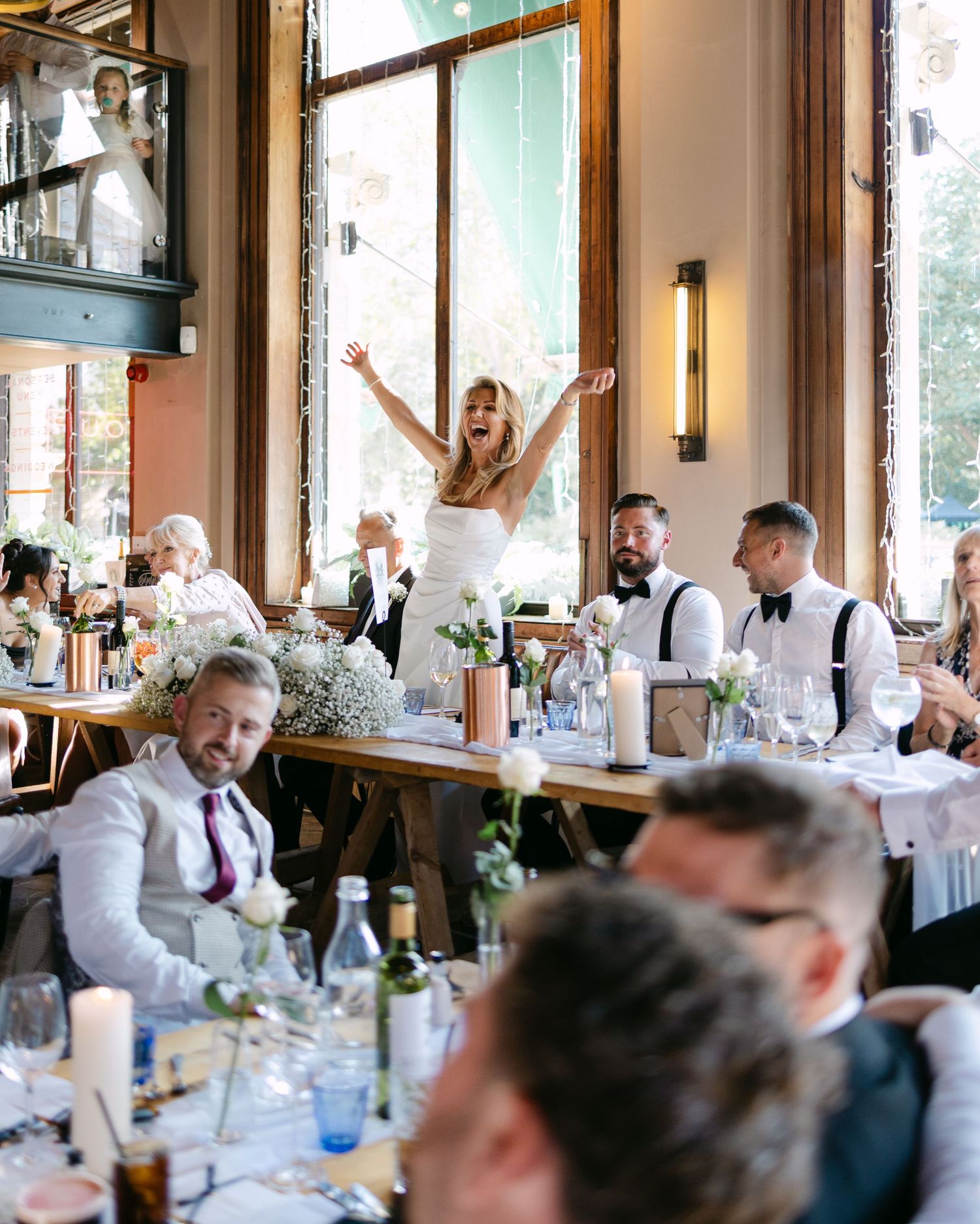 Wedding guests celebrate at a rustic reception venue with tall windows and elegant table settings.