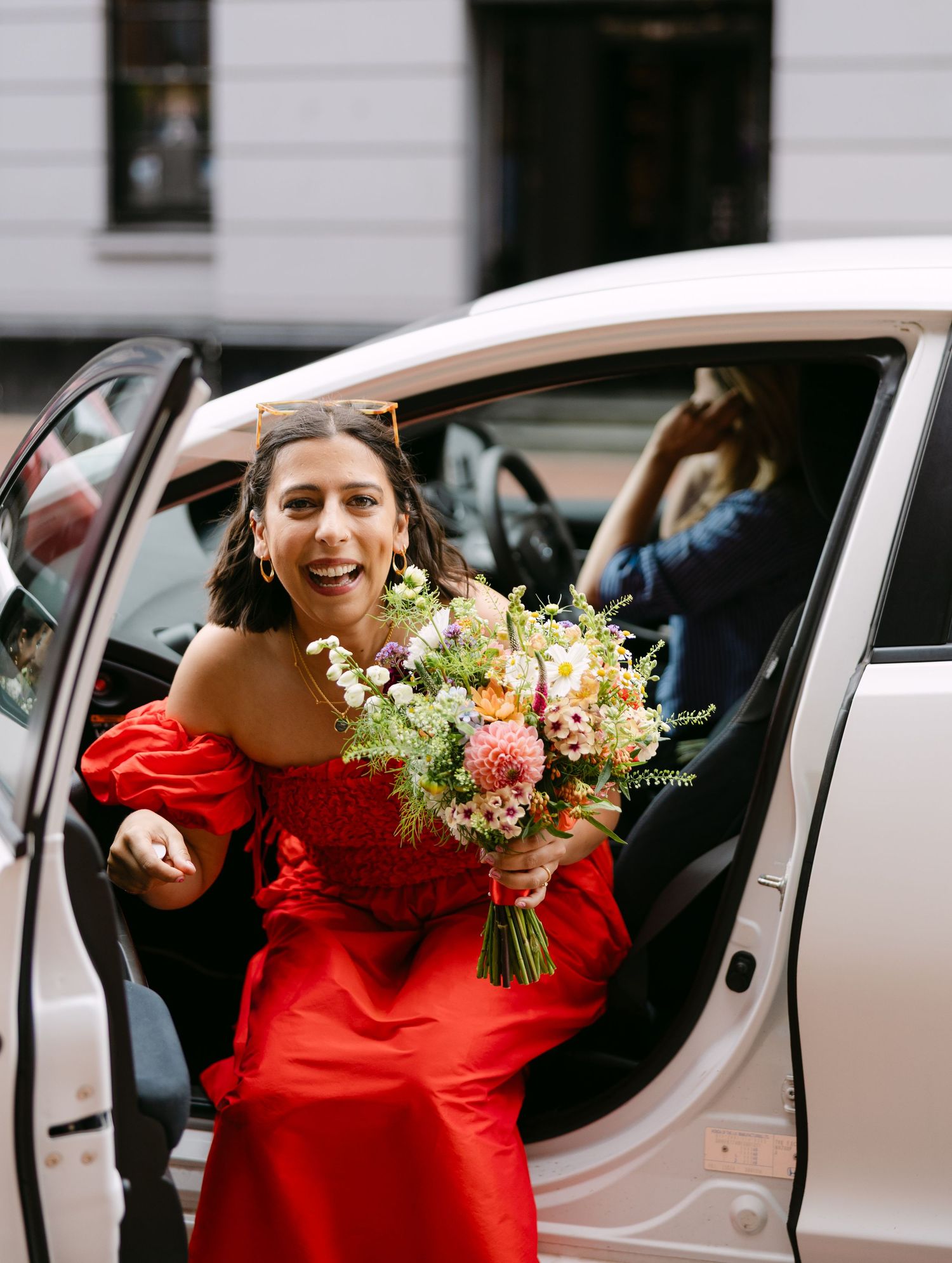 A couple in a red dress and gray suit exit a white car while holding a colorful wedding bouquet.
