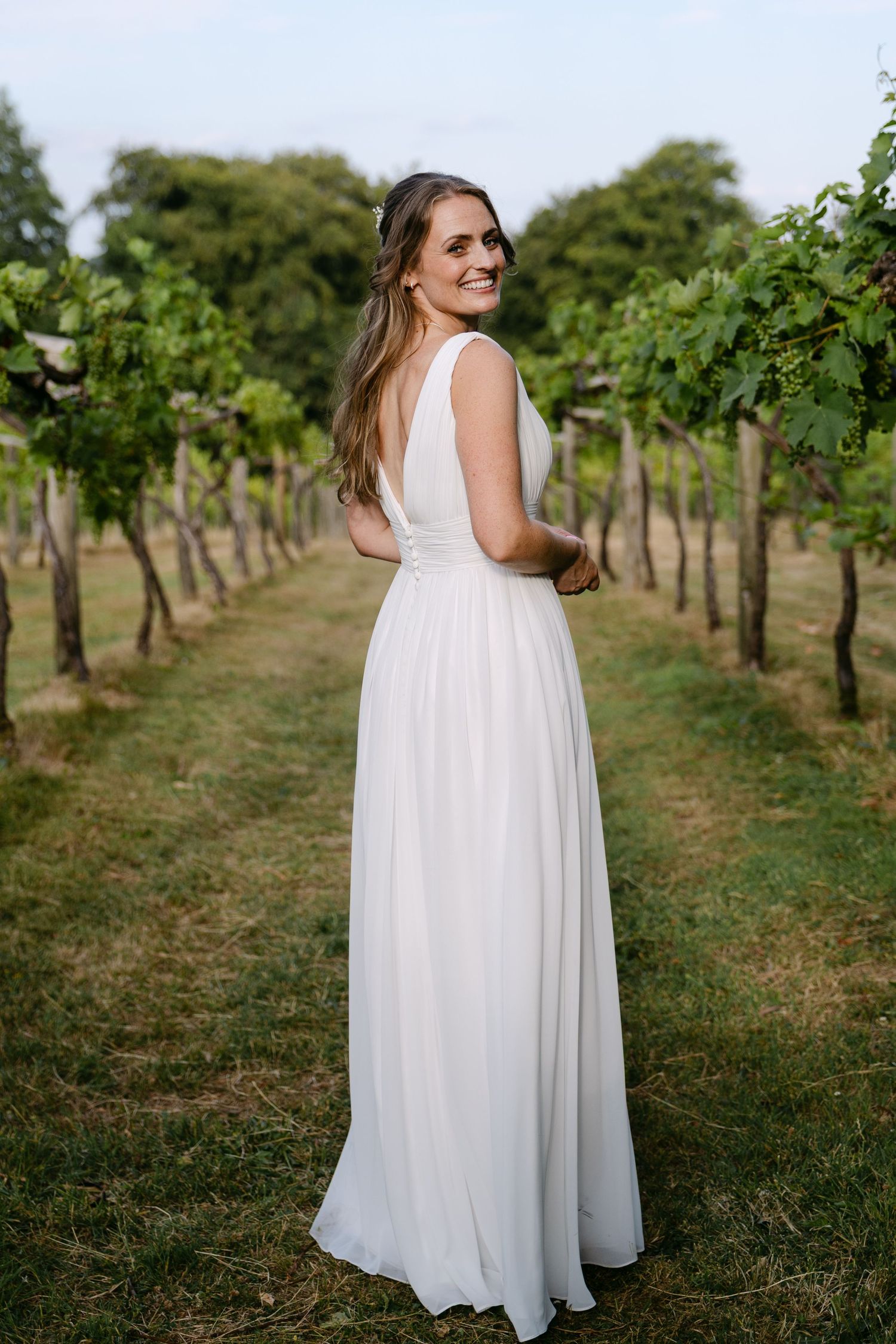 Bride in elegant white gown standing among vineyard rows, smiling over her shoulder on a lush green summer day.
