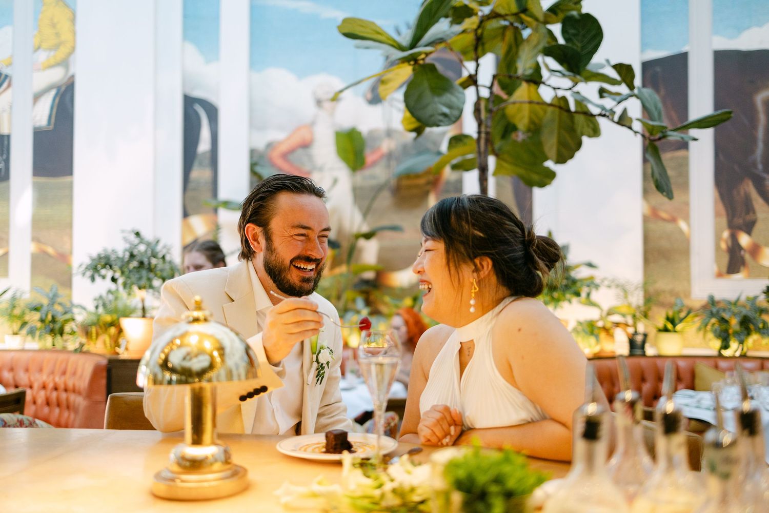 Couple laughing together at a restaurant table during their wedding reception, surrounded by lush greenery and elegant decor.