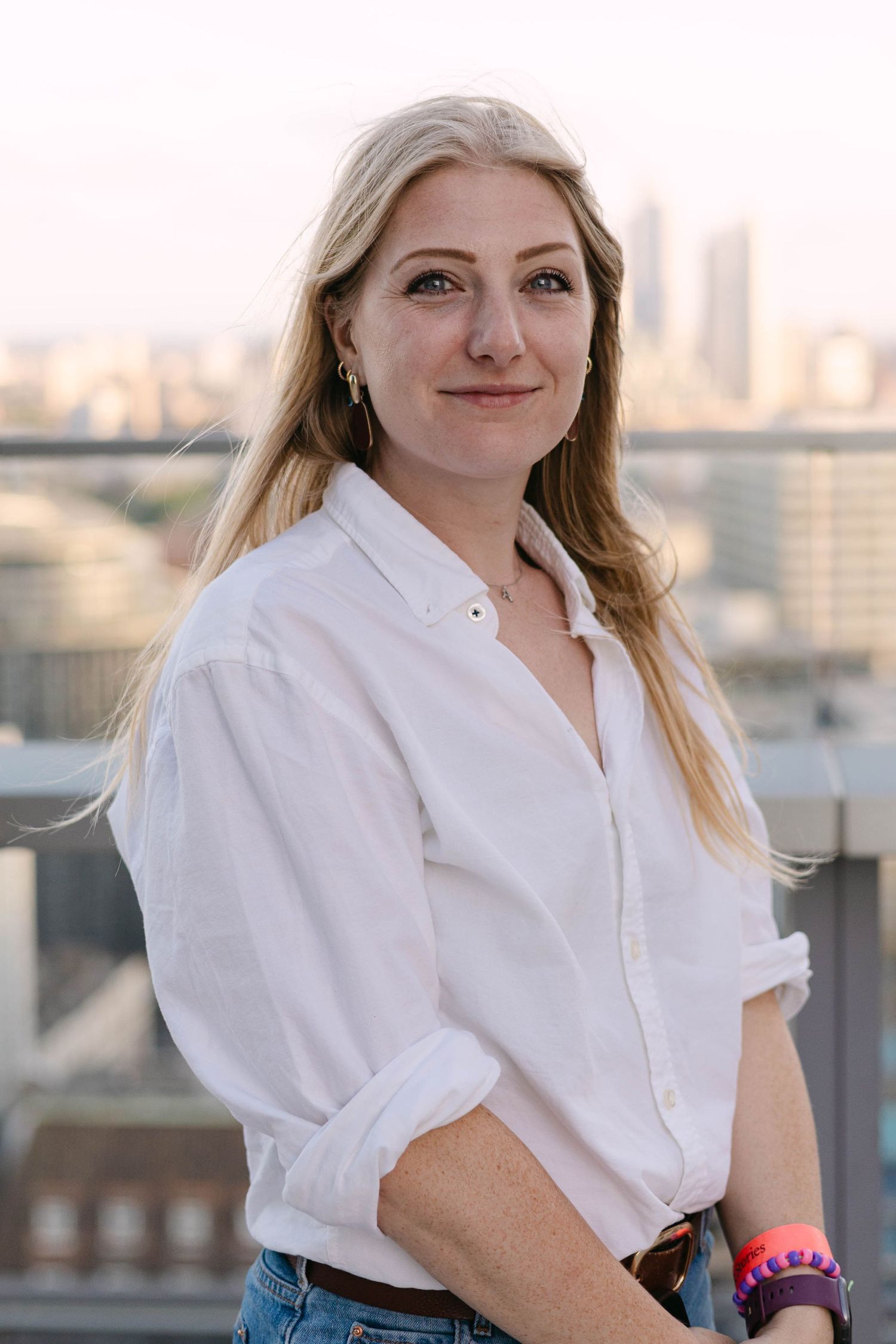 Young blonde woman in white shirt smiling on urban rooftop with city skyline at sunset.