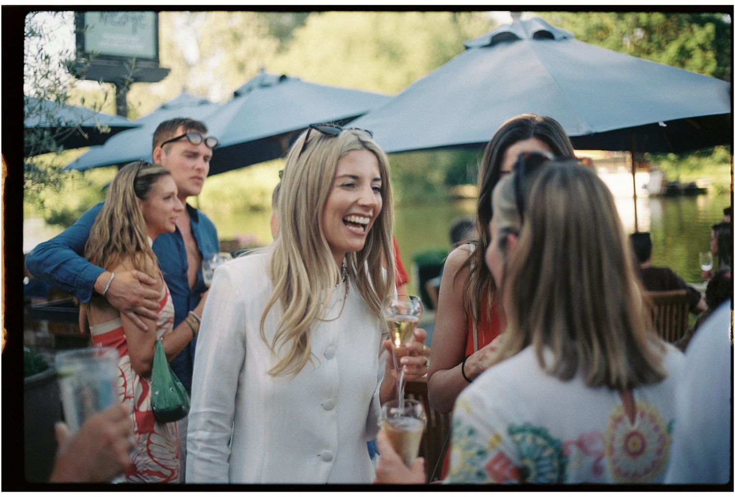 Women laughing and socializing at an outdoor garden party under blue umbrellas on a sunny day.