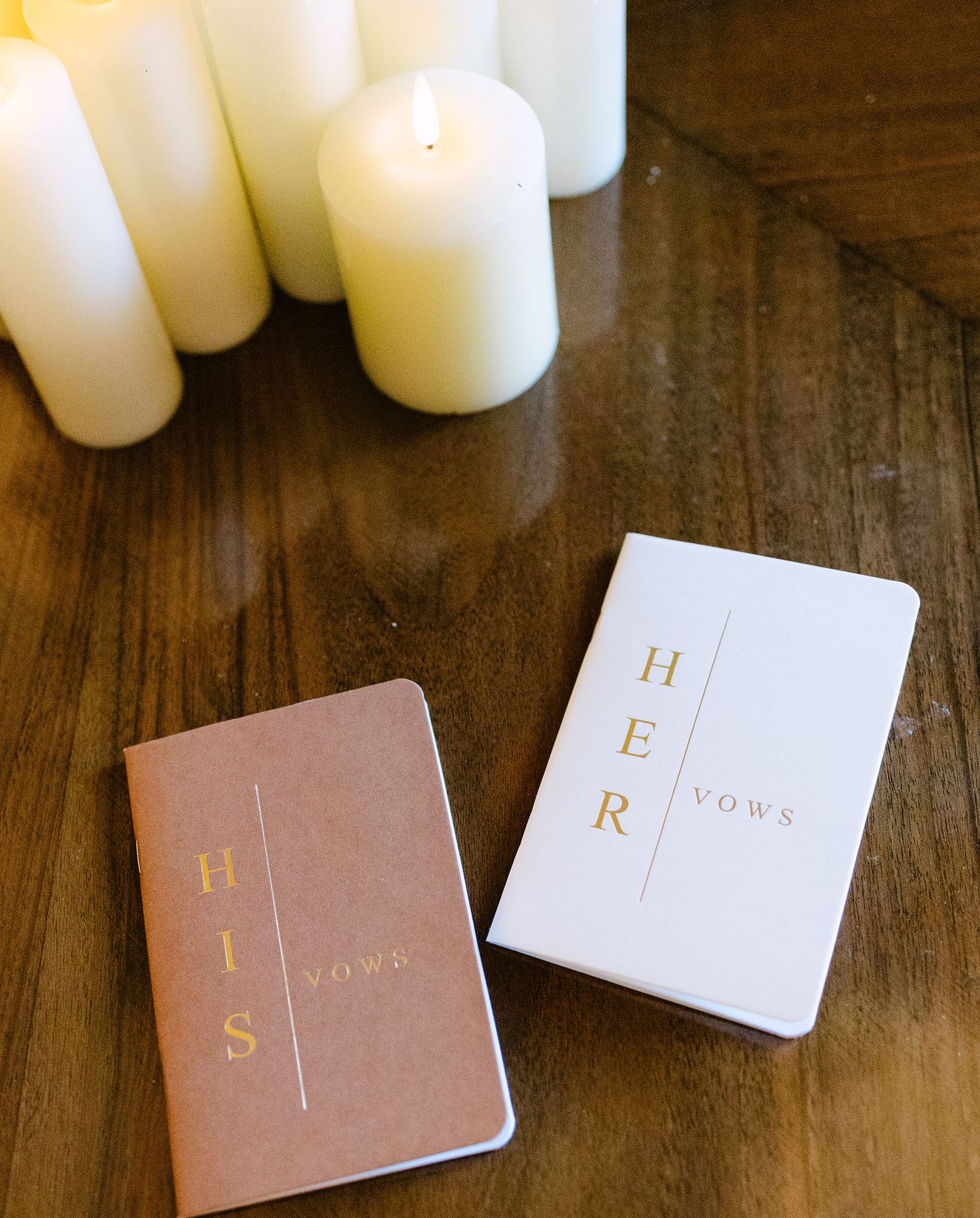 His and Her vow books with gold lettering displayed next to white candles on wooden surface.