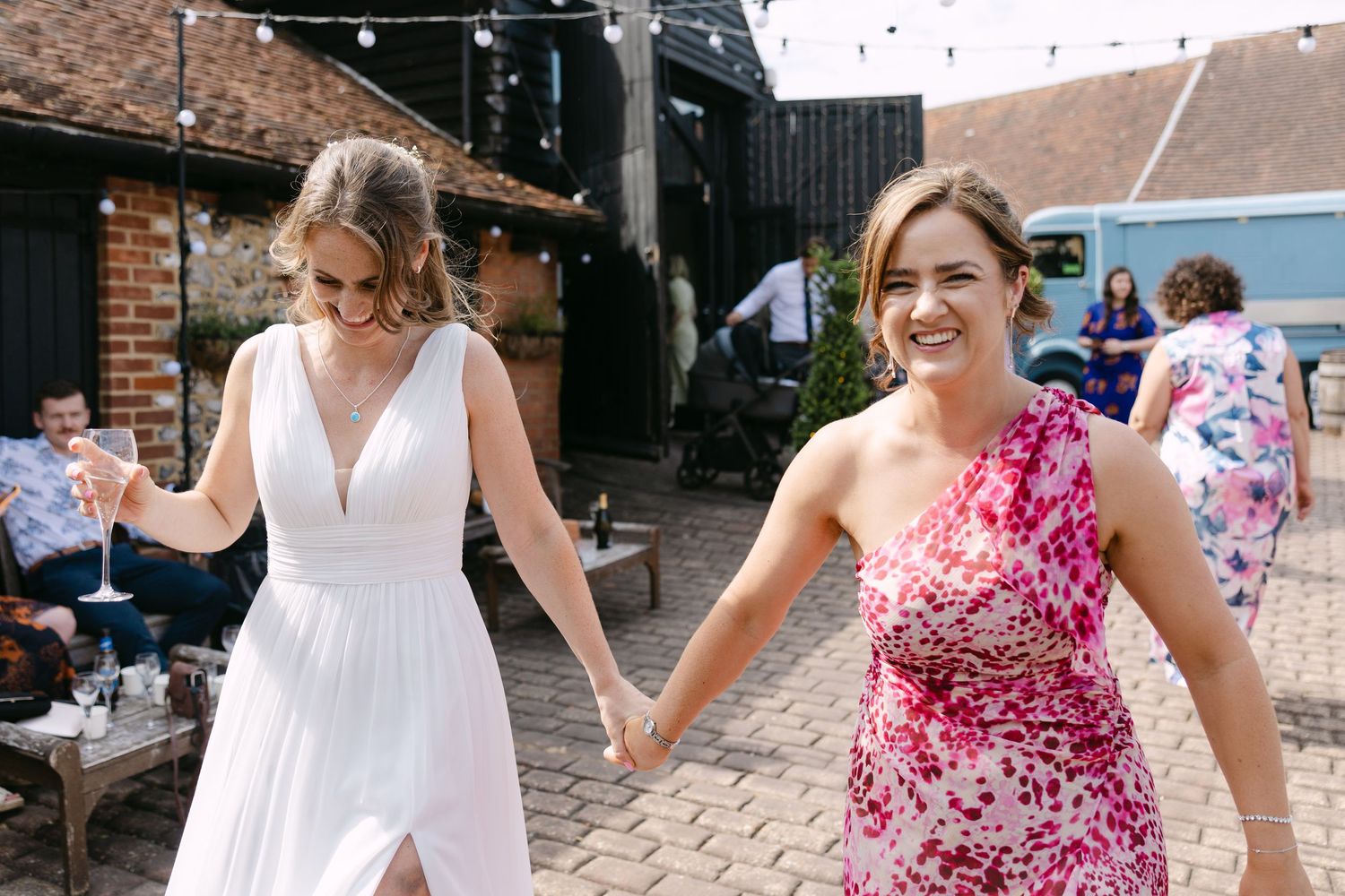Two women holding hands smiling at outdoor wedding venue, one in white bridal gown.
