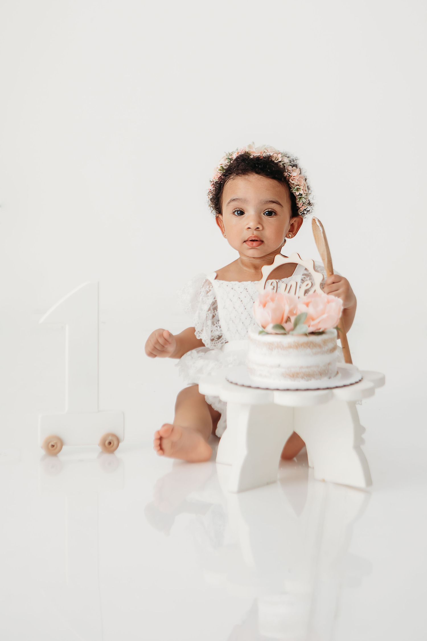 Baby in white outfit sits near birthday cake on white cake stand during first birthday cake smash photoshoot.