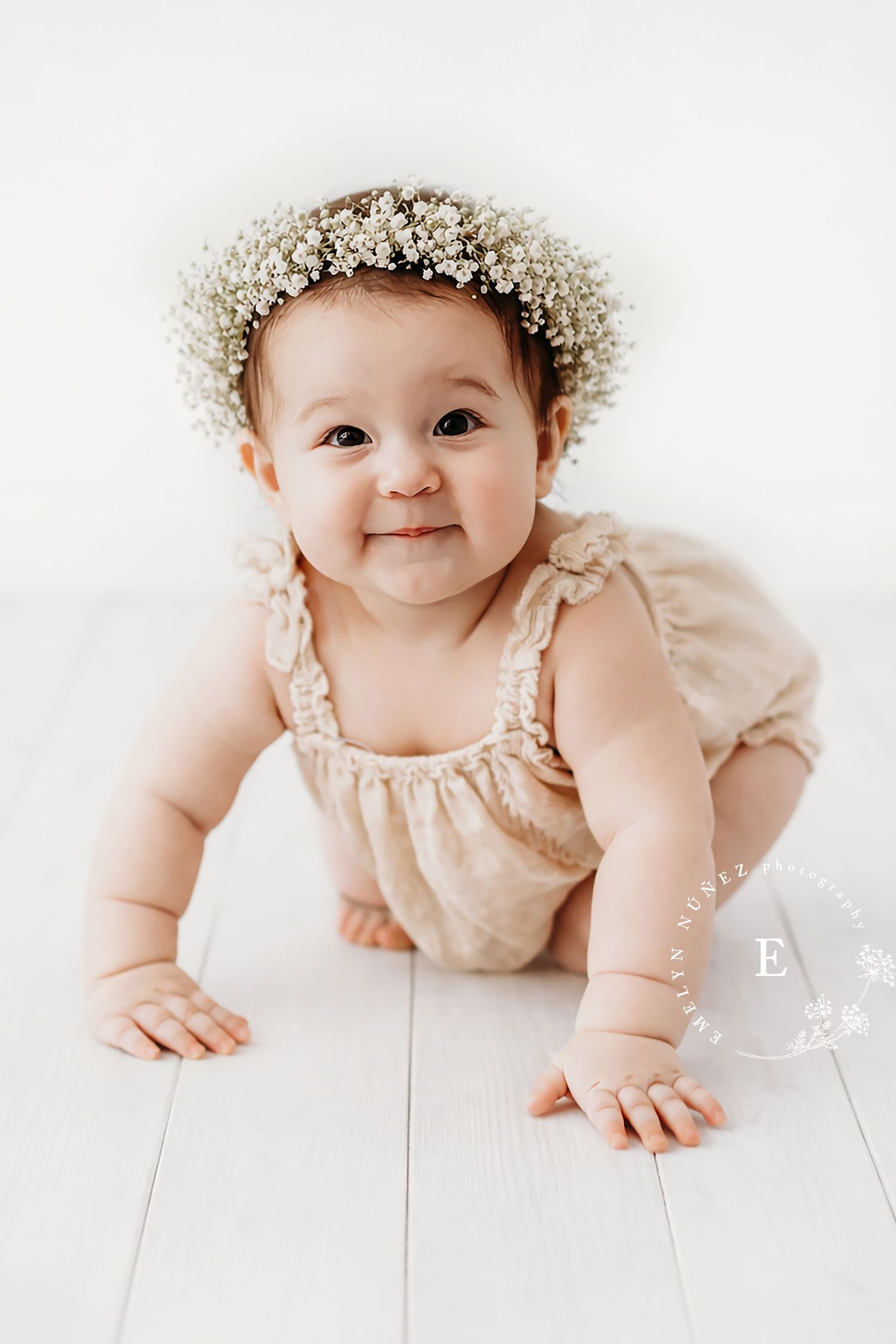 Baby wearing floral crown and beige romper crawls on white wooden floor.