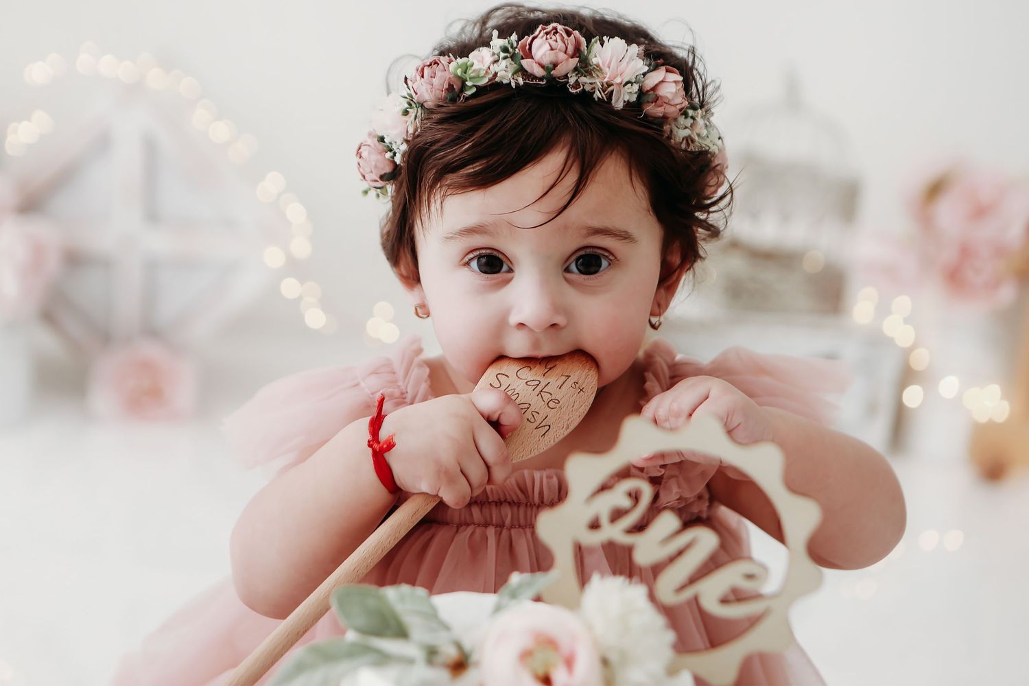 Baby in a floral crown and white dress poses against a soft white background with twinkling lights.