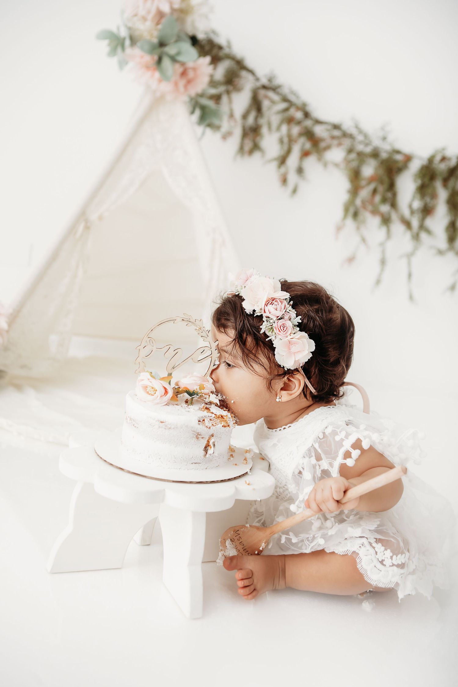 A baby in a white lace dress and floral headpiece sits on the floor eating cake during a cake smash photo session.