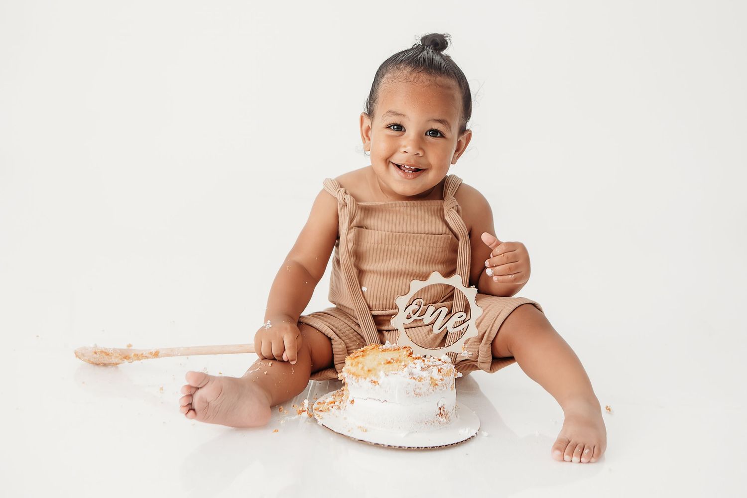 Baby in white diaper smiling while sitting next to smashed birthday cake on white background.