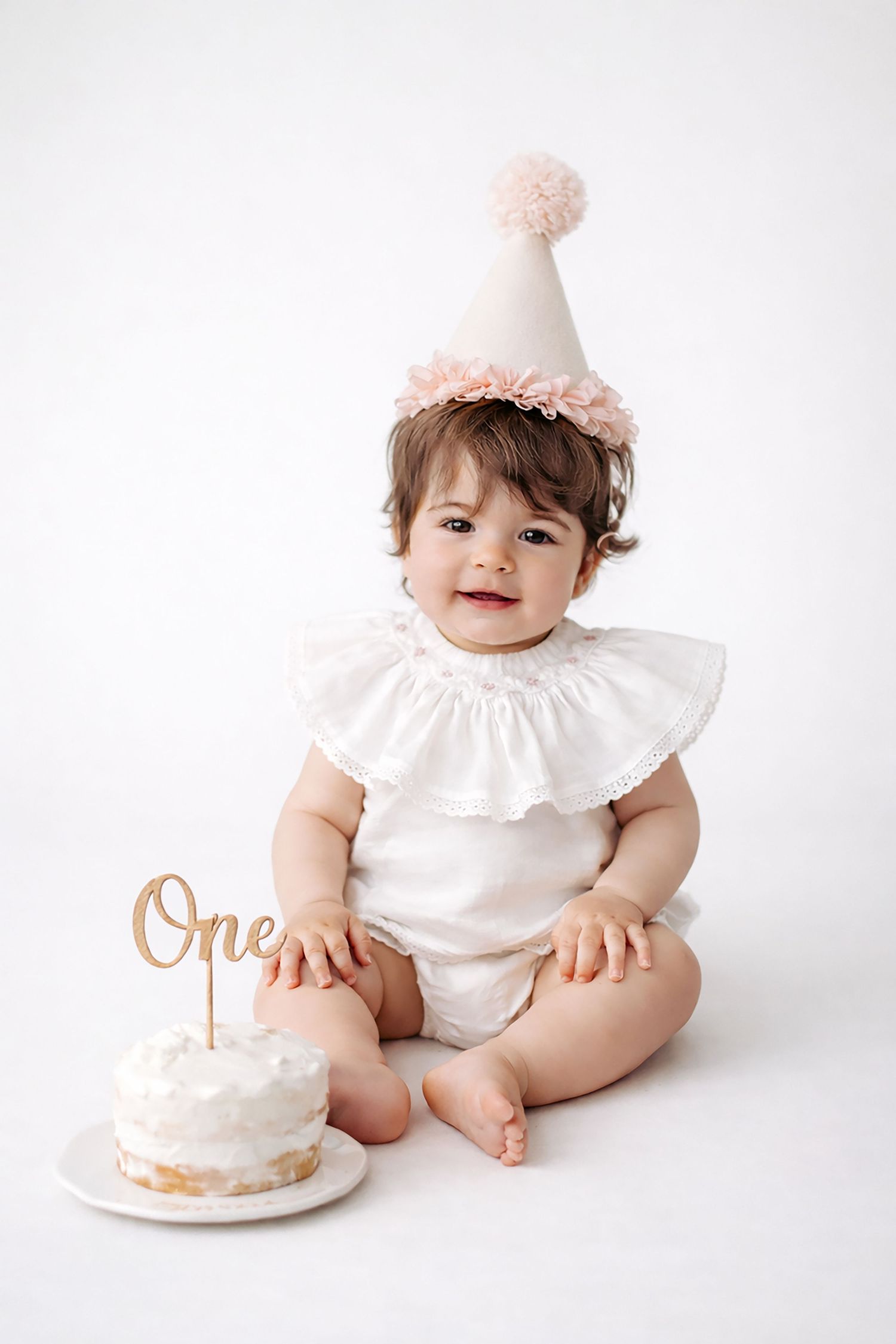 A baby in white ruffled outfit sits beside a first birthday cake during a cake smash photo session against white backdrop.