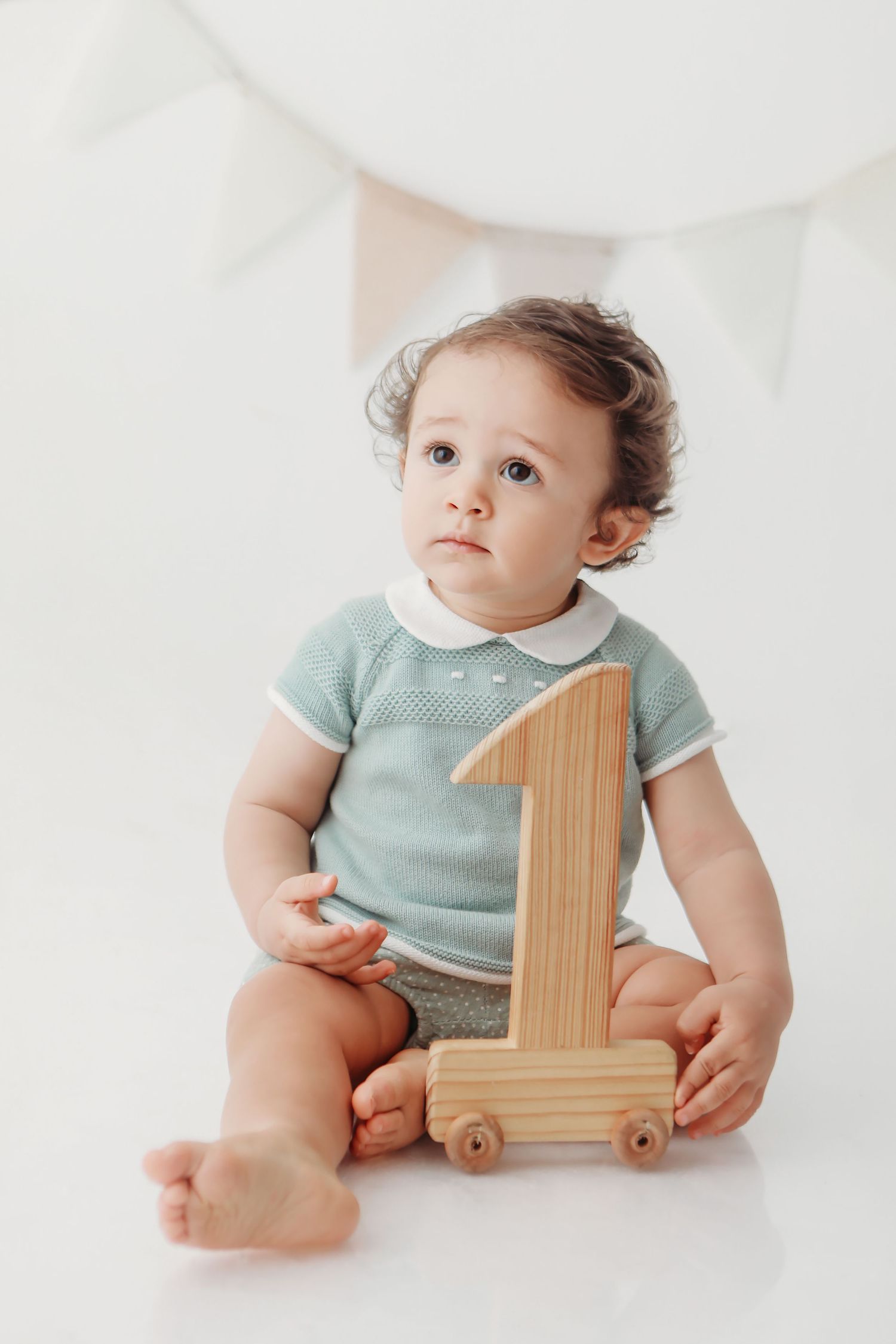 Baby in mint green outfit sits with wooden number one toy on white background.