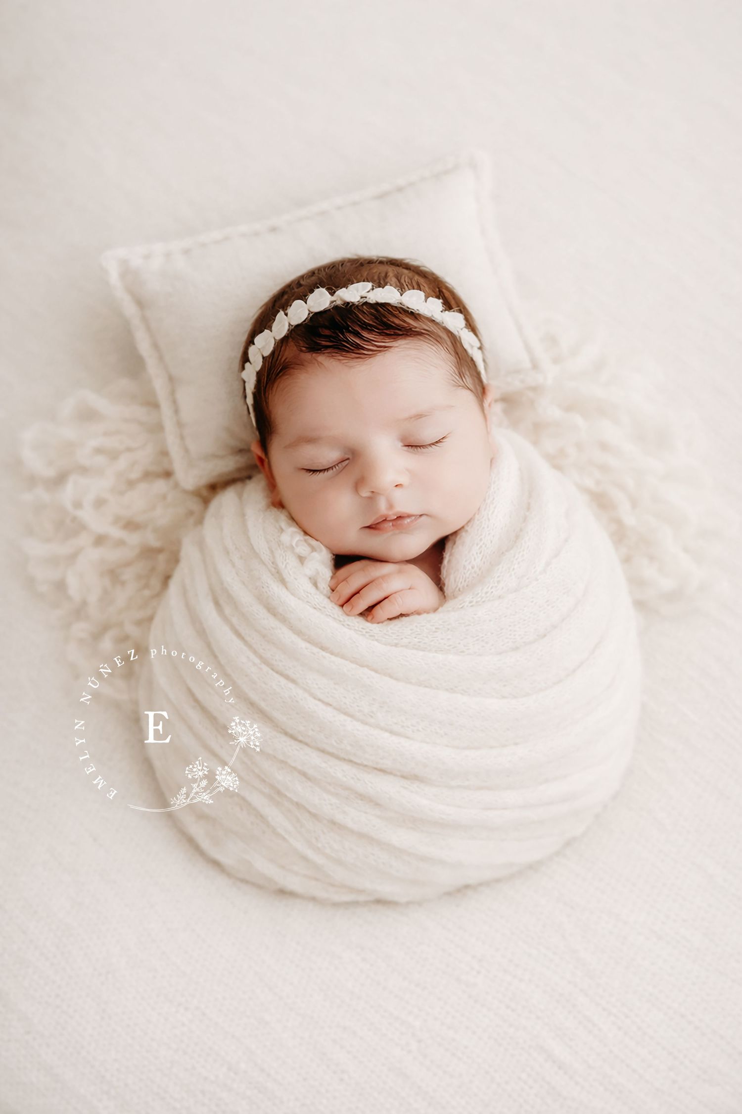 Sleeping newborn in white wrap with pearl headband on soft white pillow.