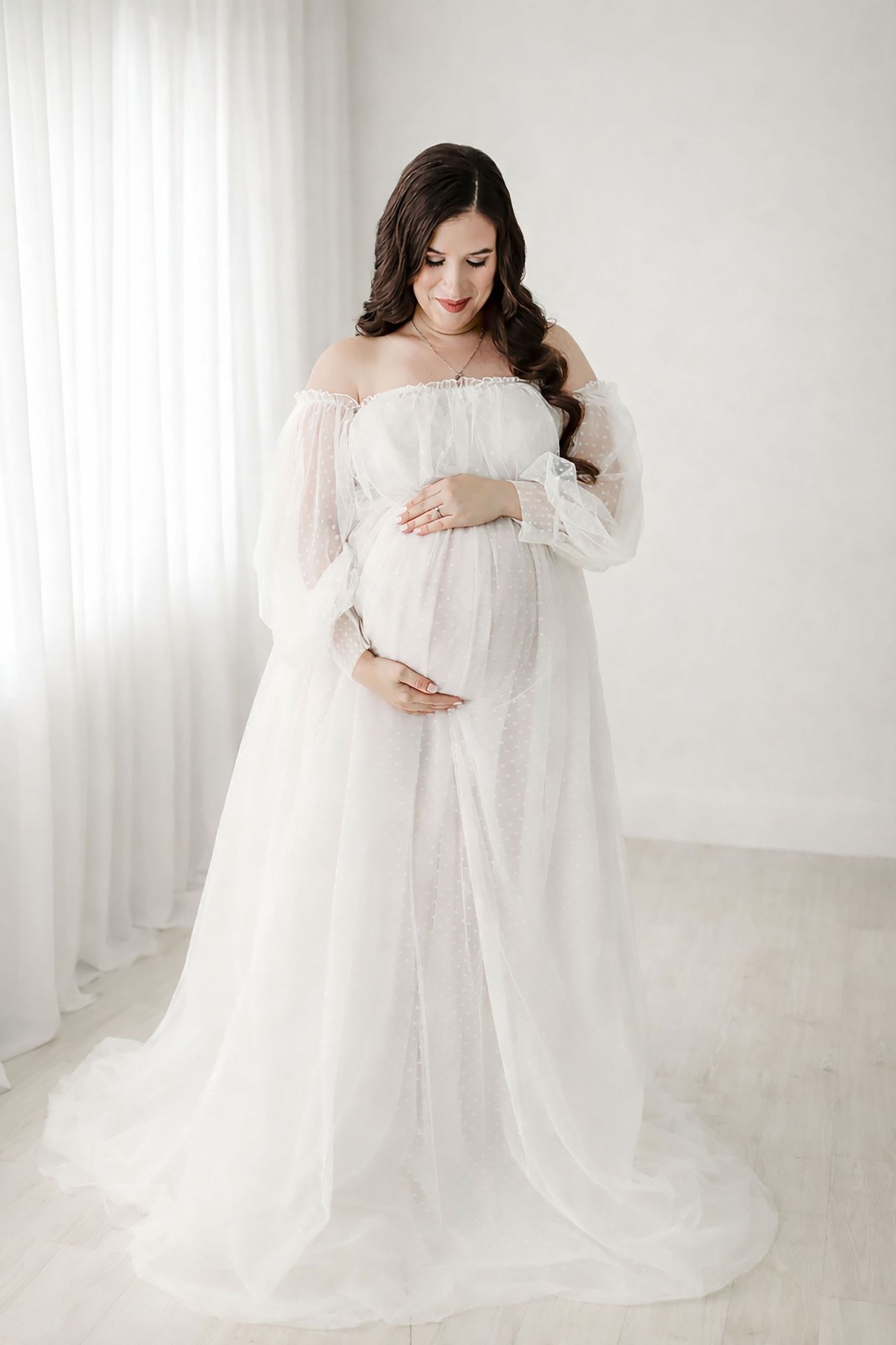 Ethereal maternity portrait in flowing white off-shoulder gown against light backdrop in minimalist setting.