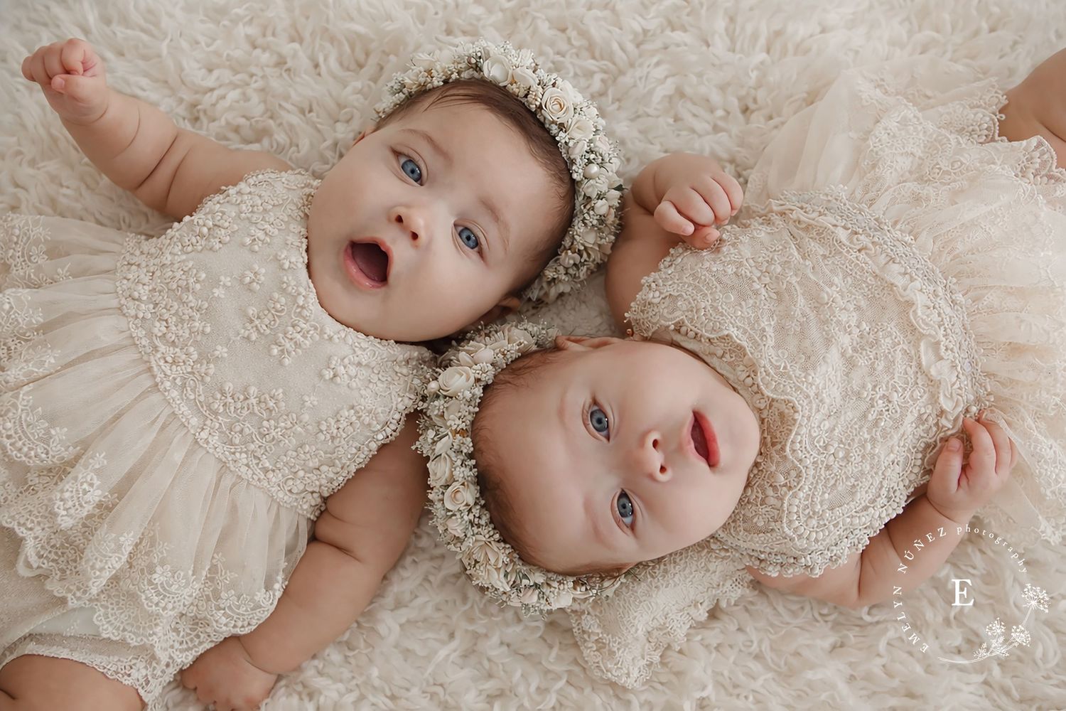 Two babies in matching lace dresses and floral crowns lying together on white background.