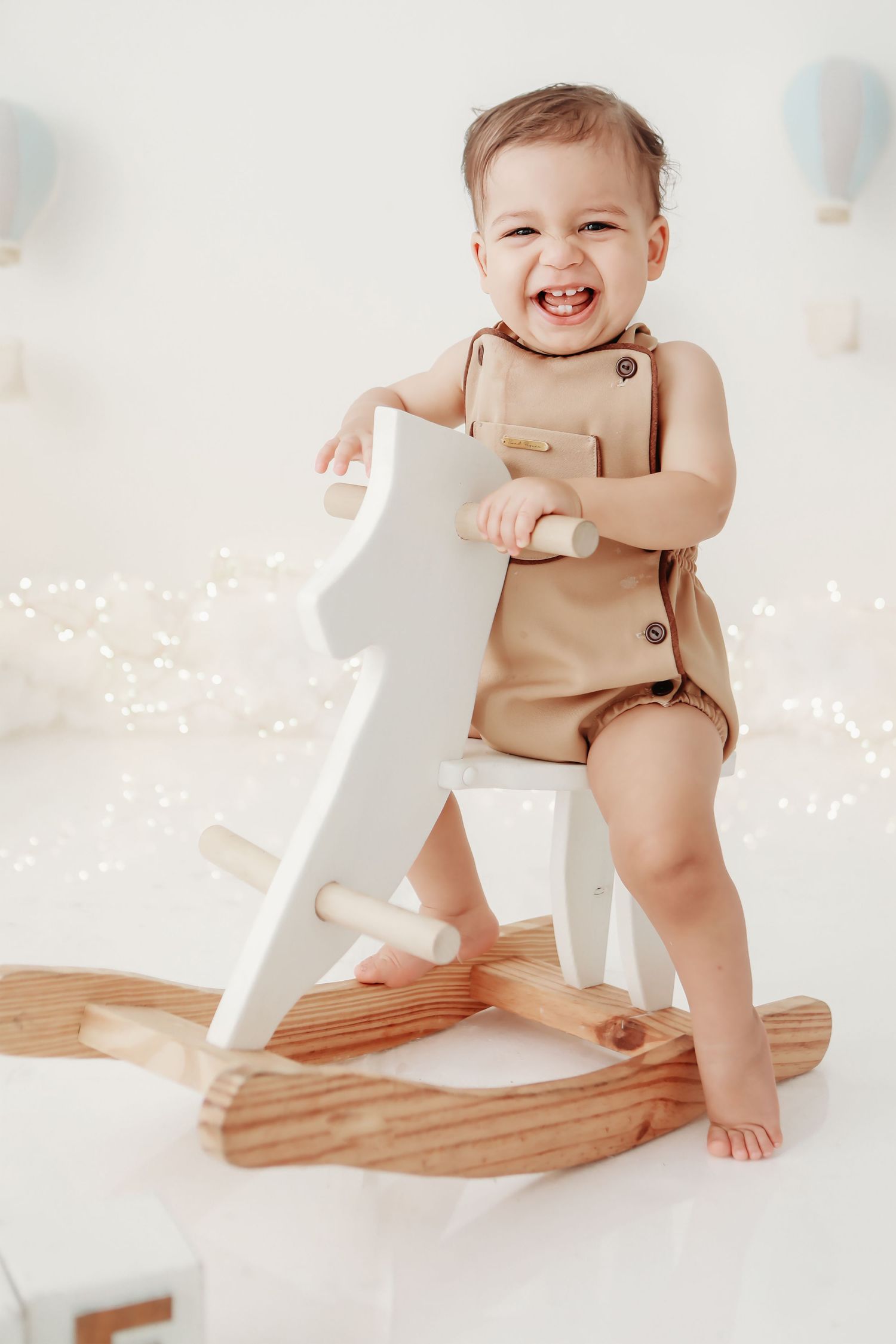 A joyful baby in beige romper plays on a wooden rocking toy against a pastel backdrop with floating balloons.