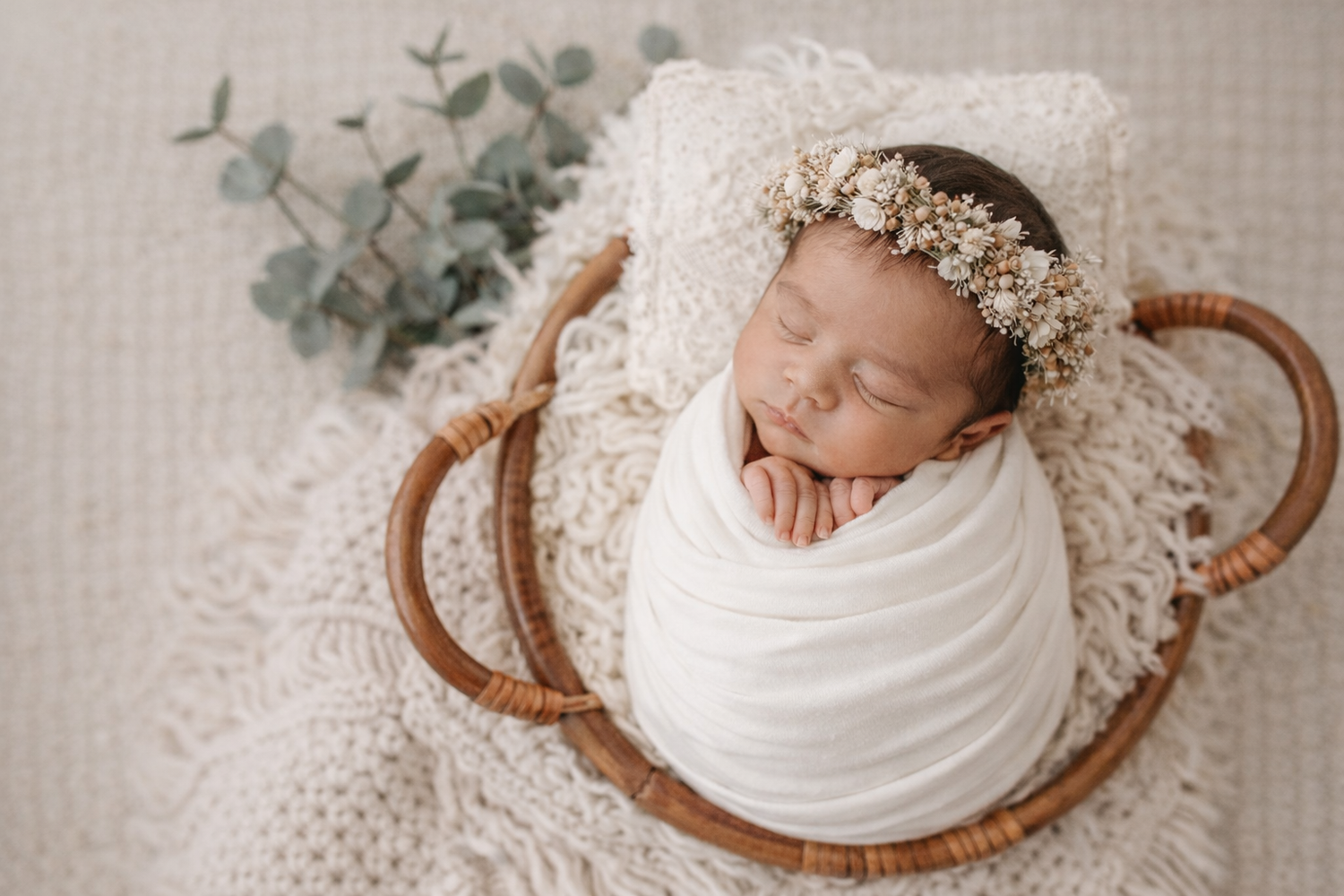 A sleeping newborn wrapped in white fabric lies in a woven basket with eucalyptus leaves and delicate floral headpiece.