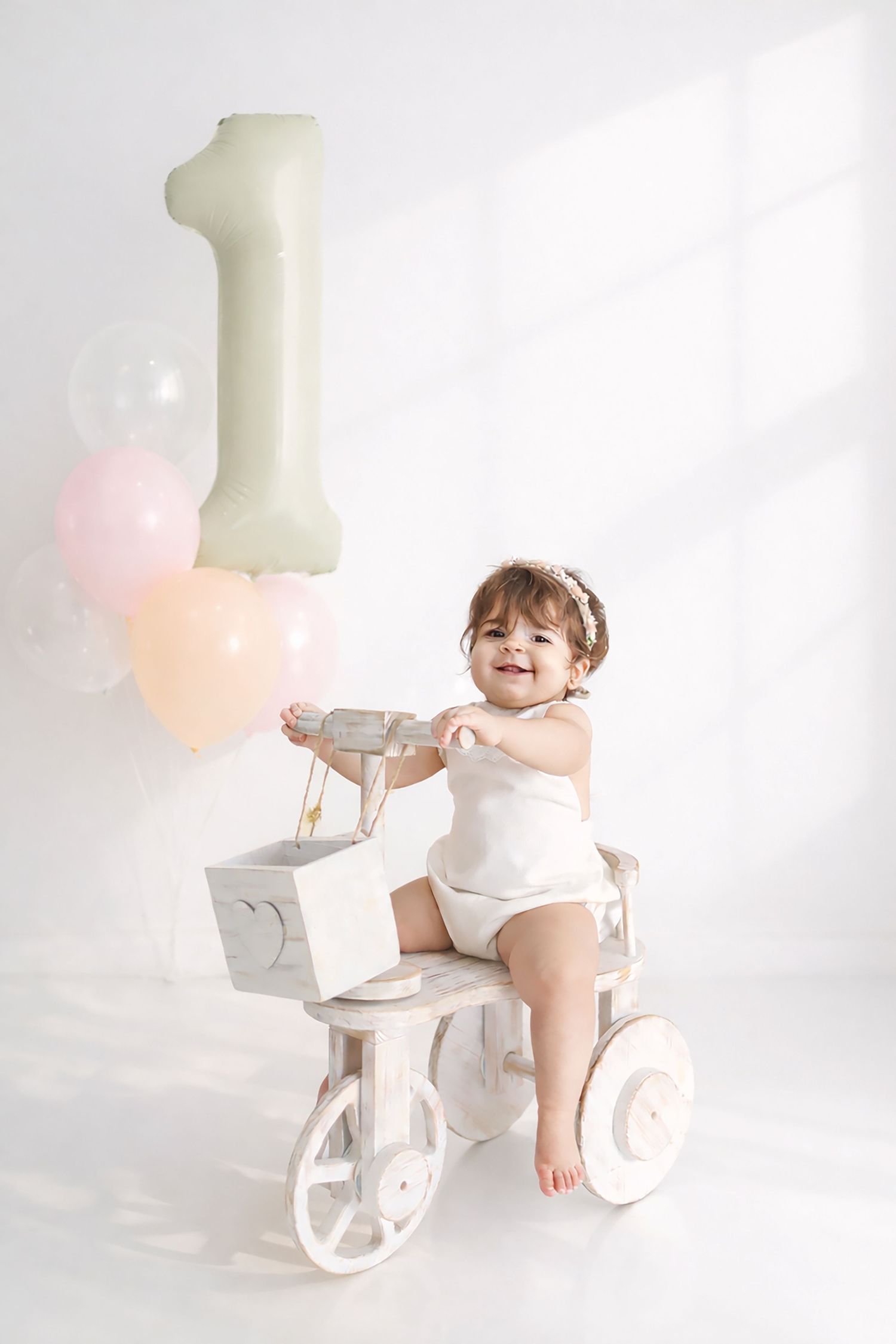 A baby in white sits on a decorative wooden tricycle with a large number 1 balloon and pastel decorations in the background.