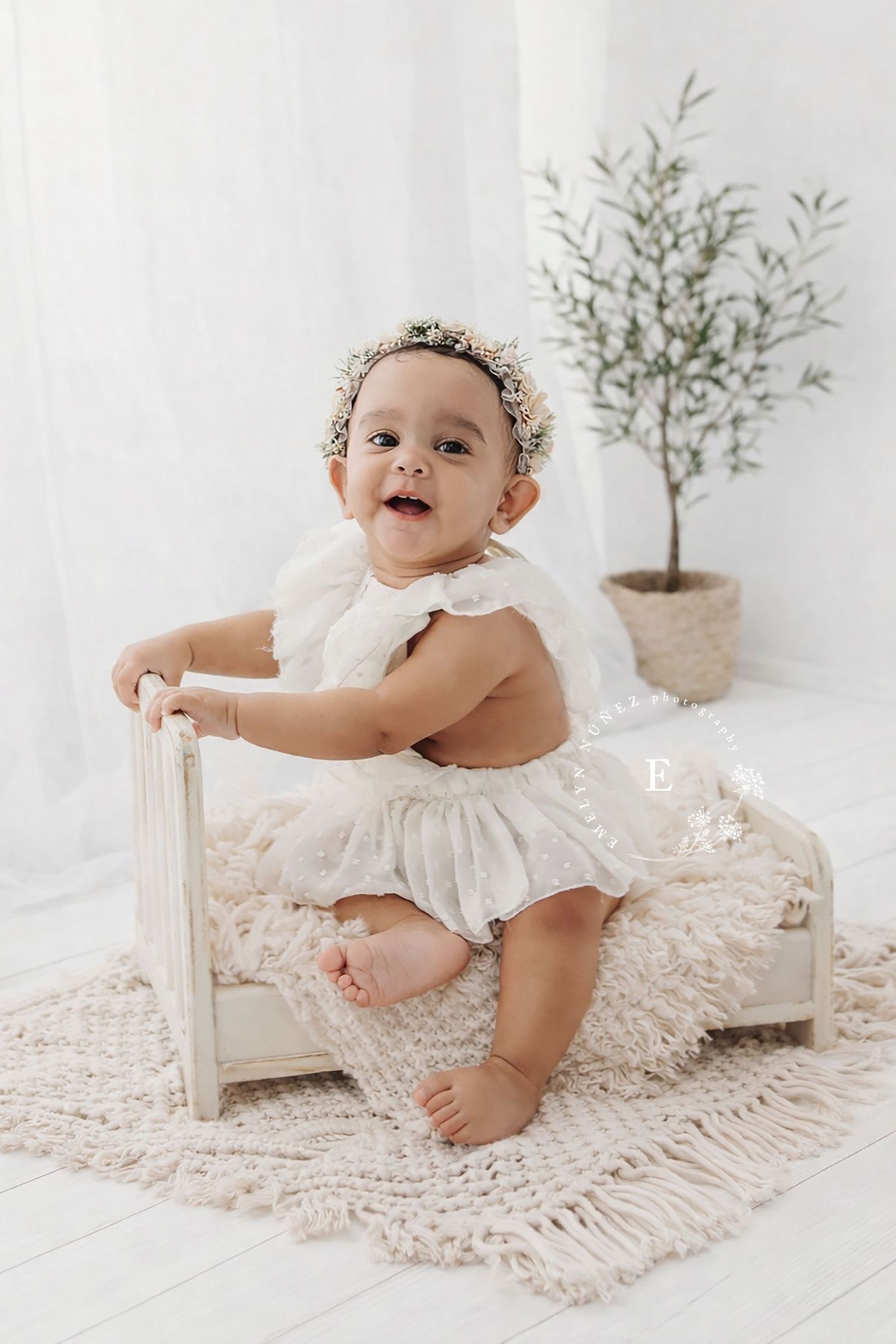 Baby in white dress sits on small chair with floral crown against white backdrop with potted plant.