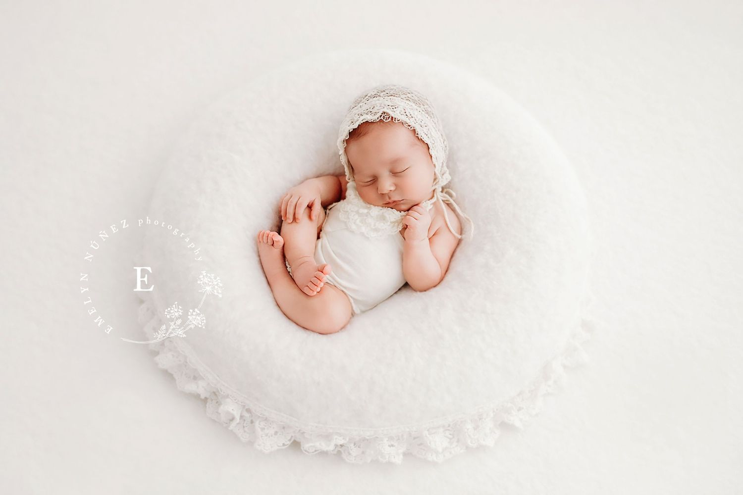 A sleeping newborn baby wrapped in soft white fabric lying on a textured white blanket in a studio setting.