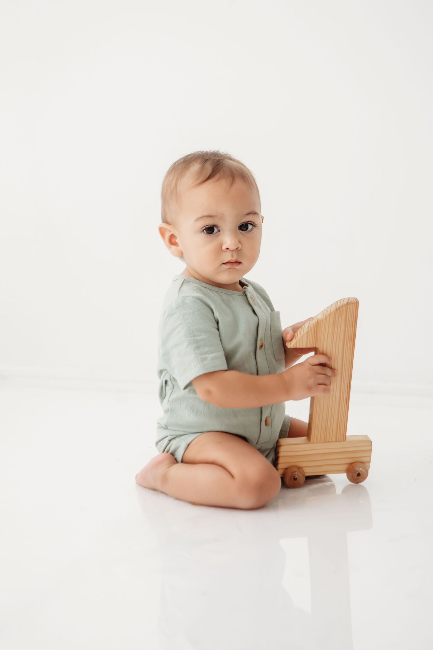 Baby wearing sage green outfit plays with wooden toy on white surface.