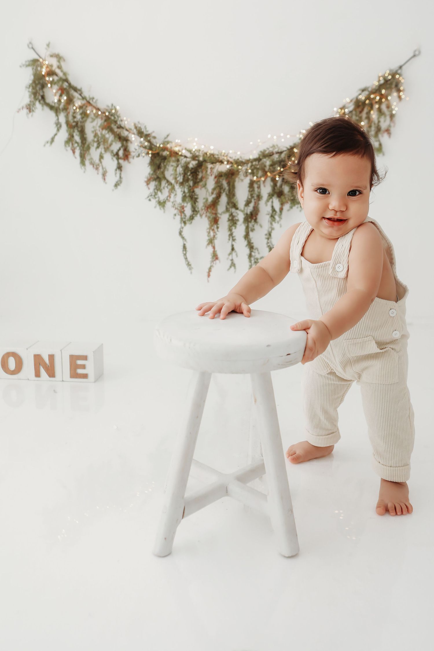Baby in cream romper plays with white chair under rustic garland decoration.