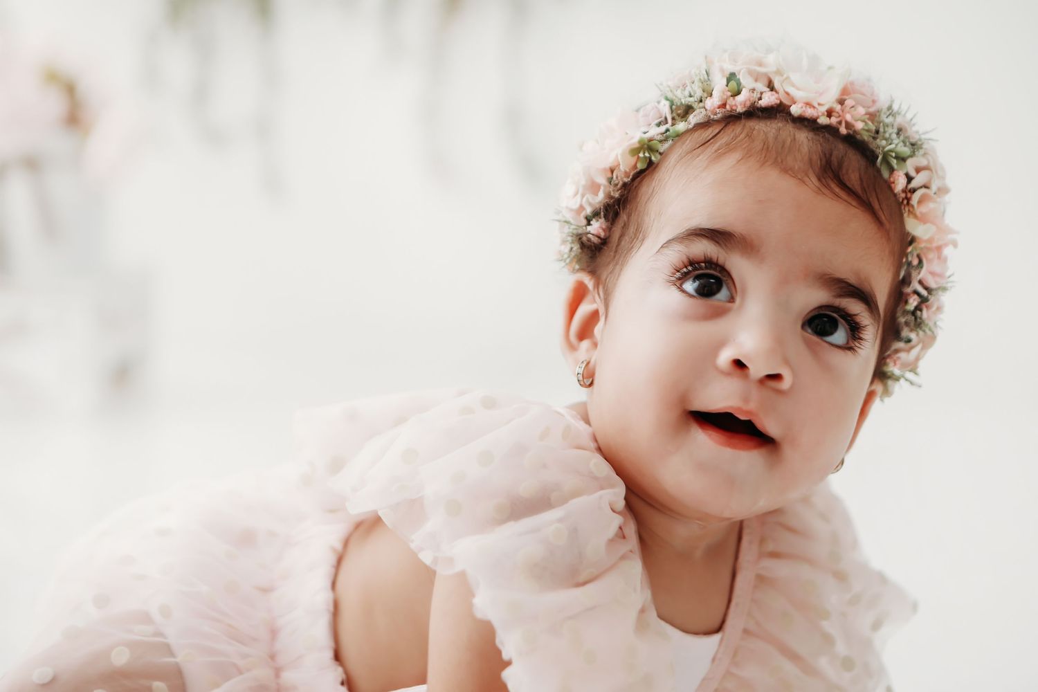 Baby wearing a delicate floral crown and lace dress smiles sweetly in a light and airy portrait.